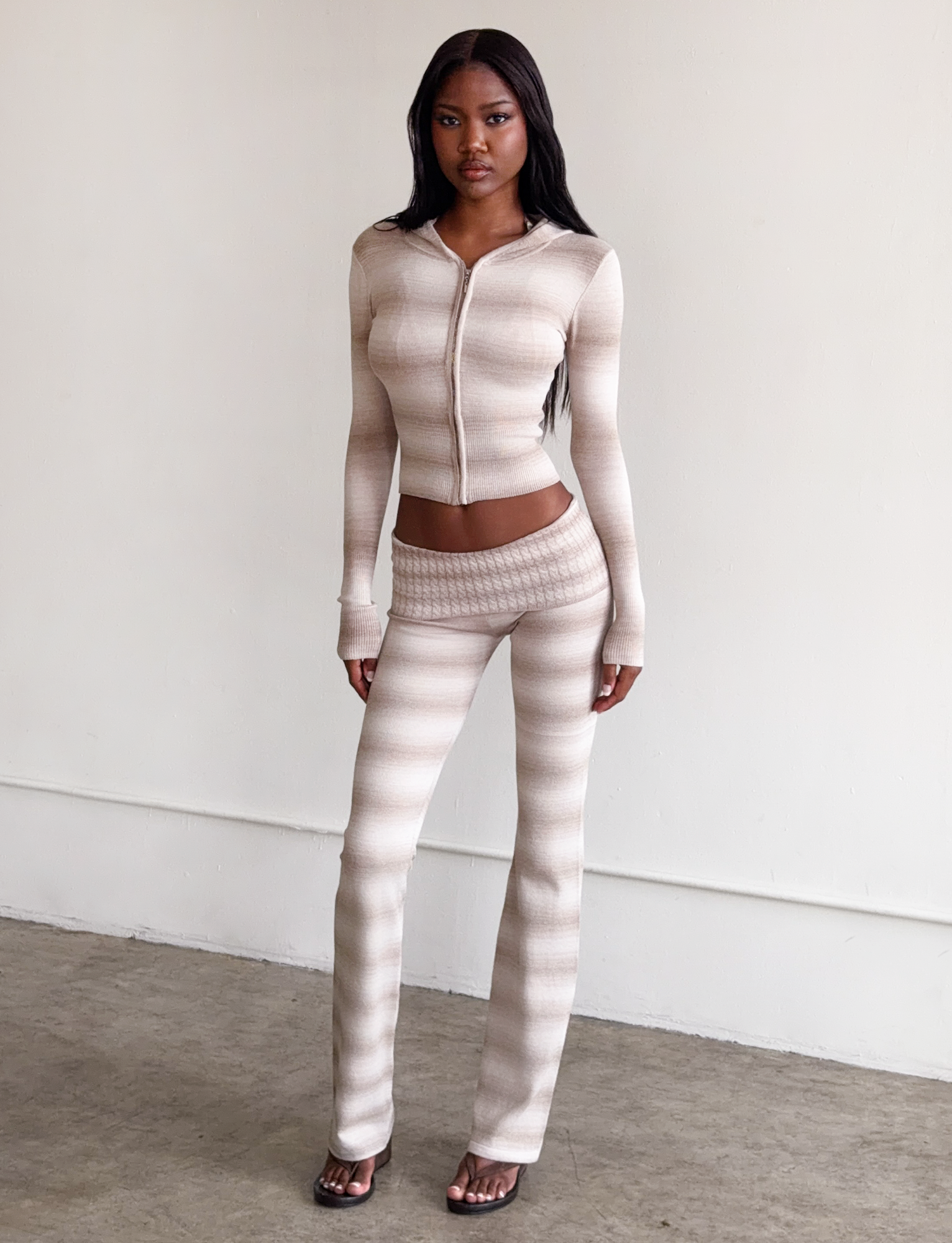 A woman with long dark hair stands indoors against a white wall, wearing TIGERMIST's MARISSA FOLDOVER KNIT PANT in taupe ombre stripe with a foldover waistband and flared leg, paired with a zip-up long-sleeve top and sandals.