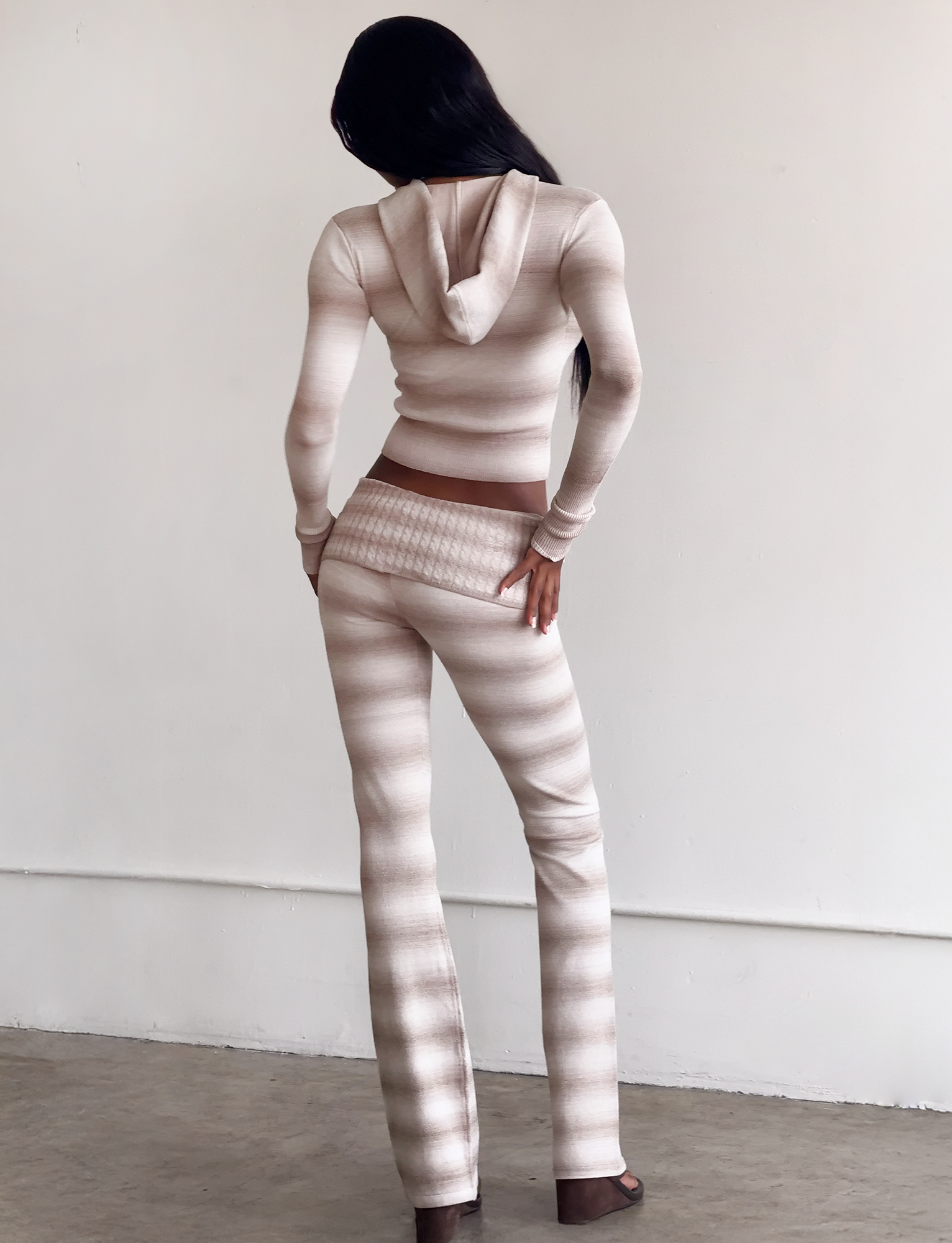 A person with long dark hair stands indoors facing away, wearing the TIGERMIST MARISSA FOLDOVER KNIT PANT in taupe ombre stripe—a fitted, hooded two-piece with a fold-over waistband and flared pants. One hand on hip; the plain background highlights the style.