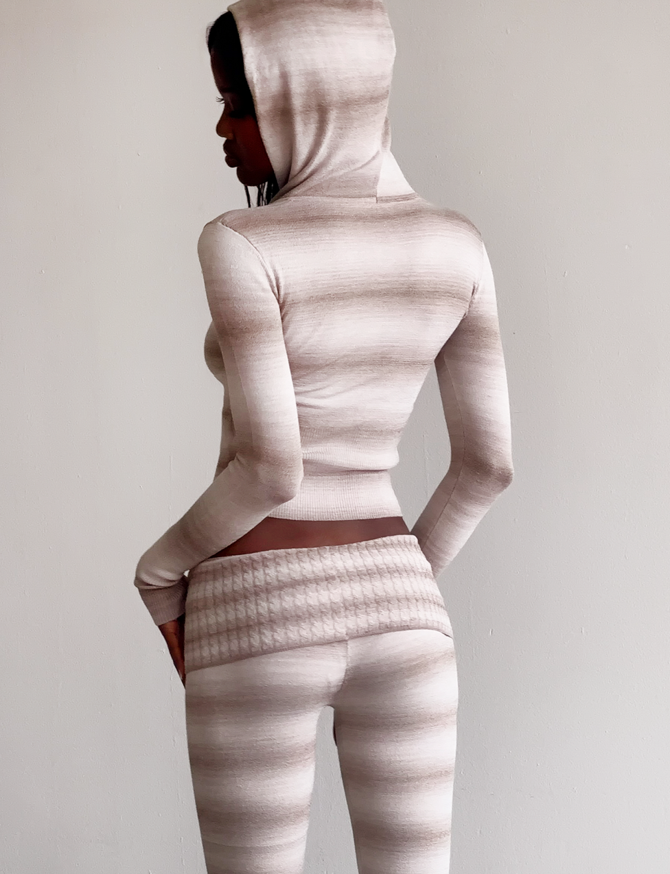 A person stands sideways, wearing the TIGERMIST MARISSA JUMPER in taupe ombre stripe with matching pants, against a plain light-colored background.