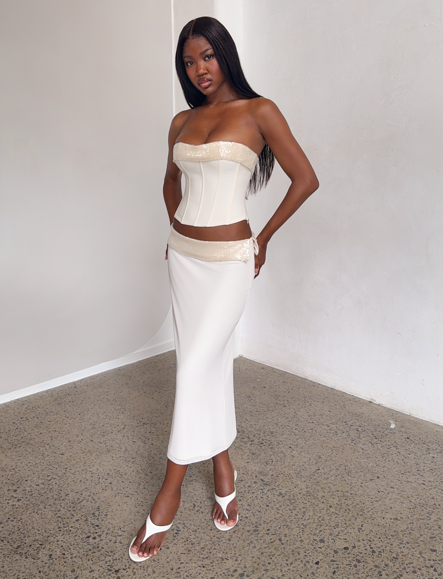 A woman poses confidently indoors in the TIGERMIST CHARLYZE MIDI SKIRT - CREAM, a cream-colored, sequin-detailed skirt, paired with a matching strapless corset top and white heeled sandals against a minimalist backdrop.