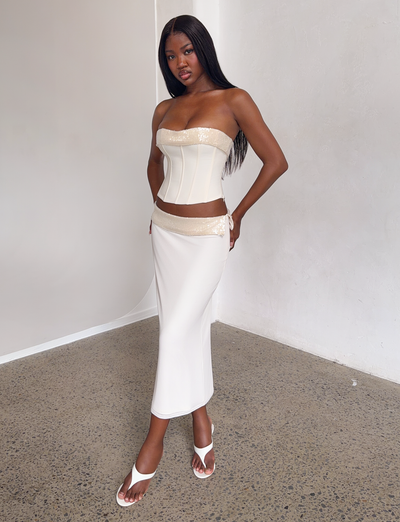A woman poses confidently indoors in the TIGERMIST CHARLYZE MIDI SKIRT - CREAM, a cream-colored, sequin-detailed skirt, paired with a matching strapless corset top and white heeled sandals against a minimalist backdrop.
