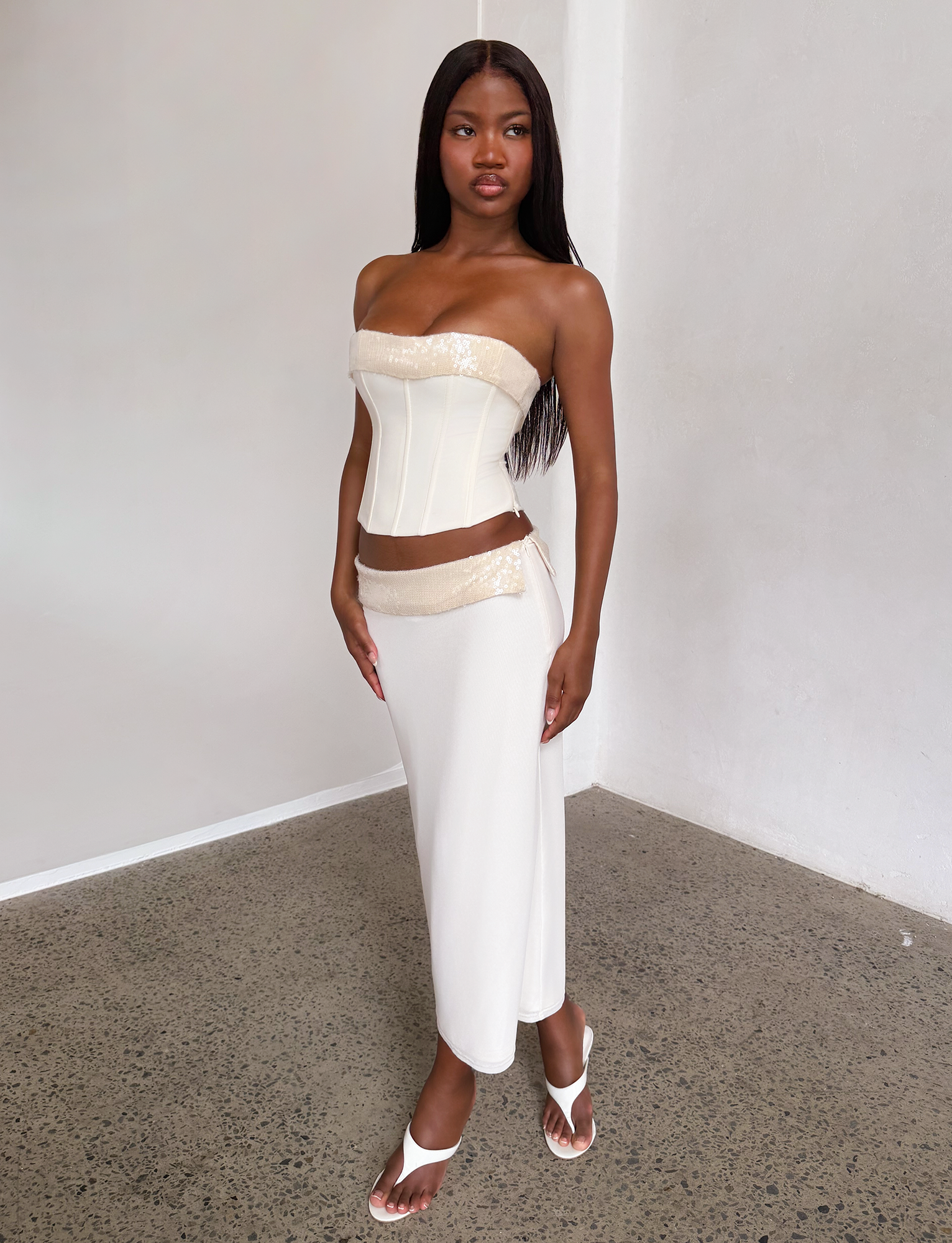 A woman poses confidently indoors against a white wall, wearing the TIGERMIST CHARLYZE MIDI SKIRT in cream with gold accents, a matching strapless corset top, and white heeled sandals. Her long hair is styled straight.