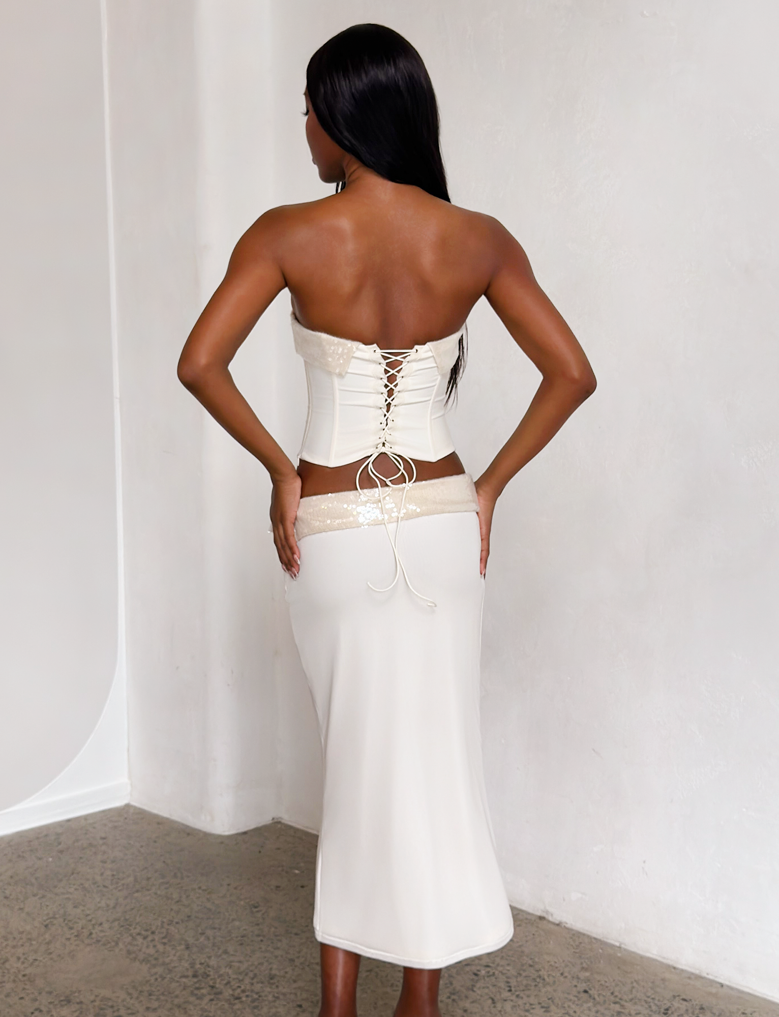A woman faces away from the camera, wearing the TIGERMIST CHARLYZE MIDI SKIRT in cream—a body-hugging skirt with lace details and a sequin waistband—paired with a white strapless lace-up corset top against a plain light wall.