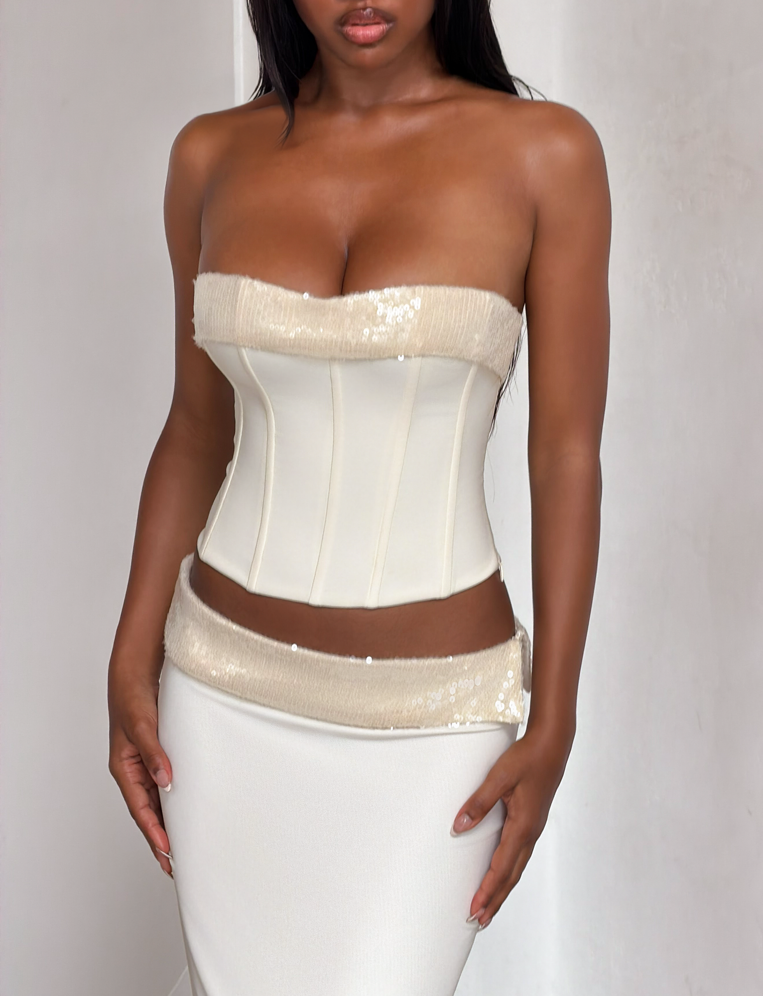 A woman wears the TIGERMIST CHARLYZE BUSTIER in cream, featuring a strapless design with sequin panels and a sweetheart neckline, paired with a matching high-waisted skirt with a sequined waistband, standing against a plain background.