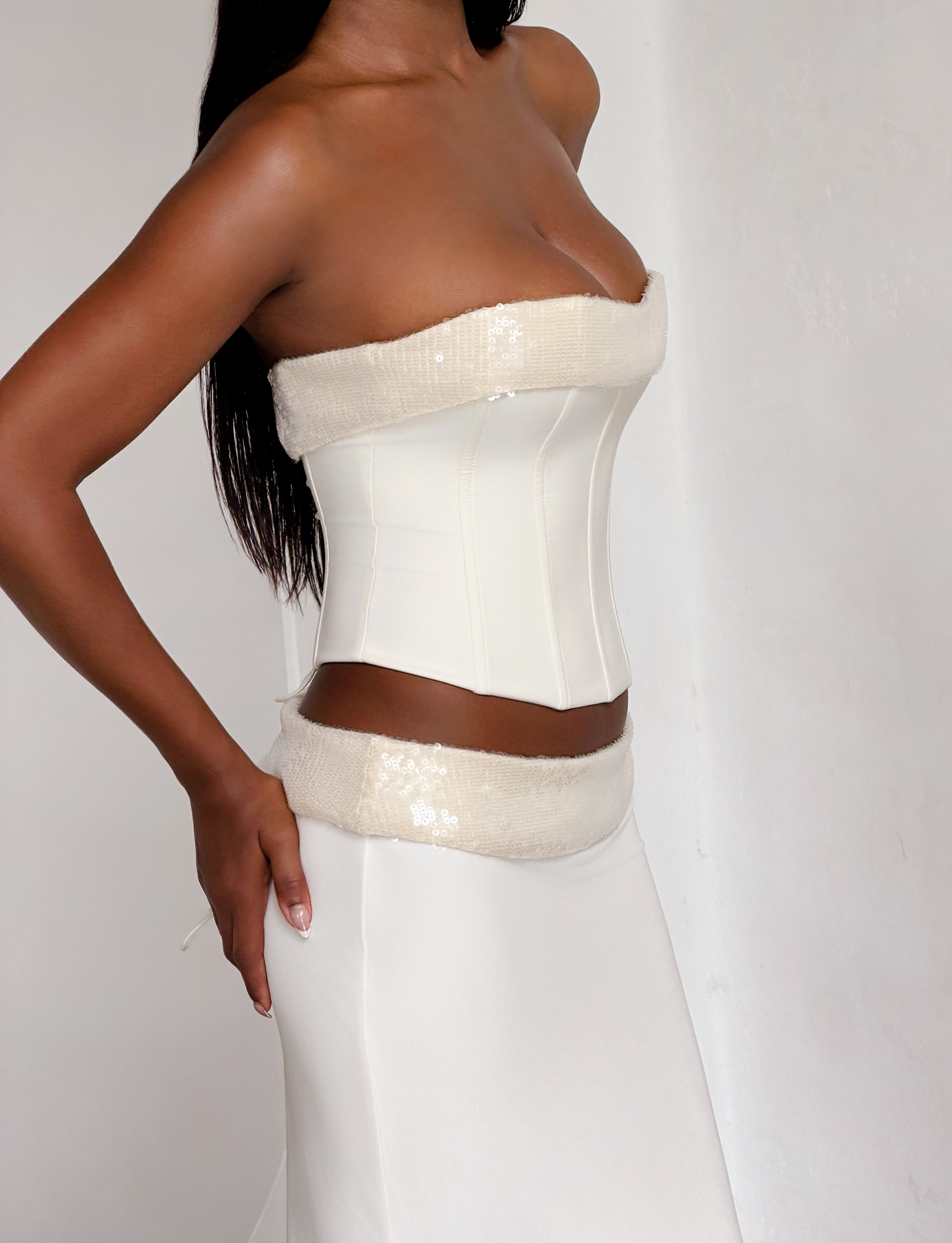 A woman stands against a plain white wall wearing the TIGERMIST CHARLYZE BUSTIER in cream, a strapless bustier top with a sequin panel, paired with a matching long white skirt. Her head is out of the frame.