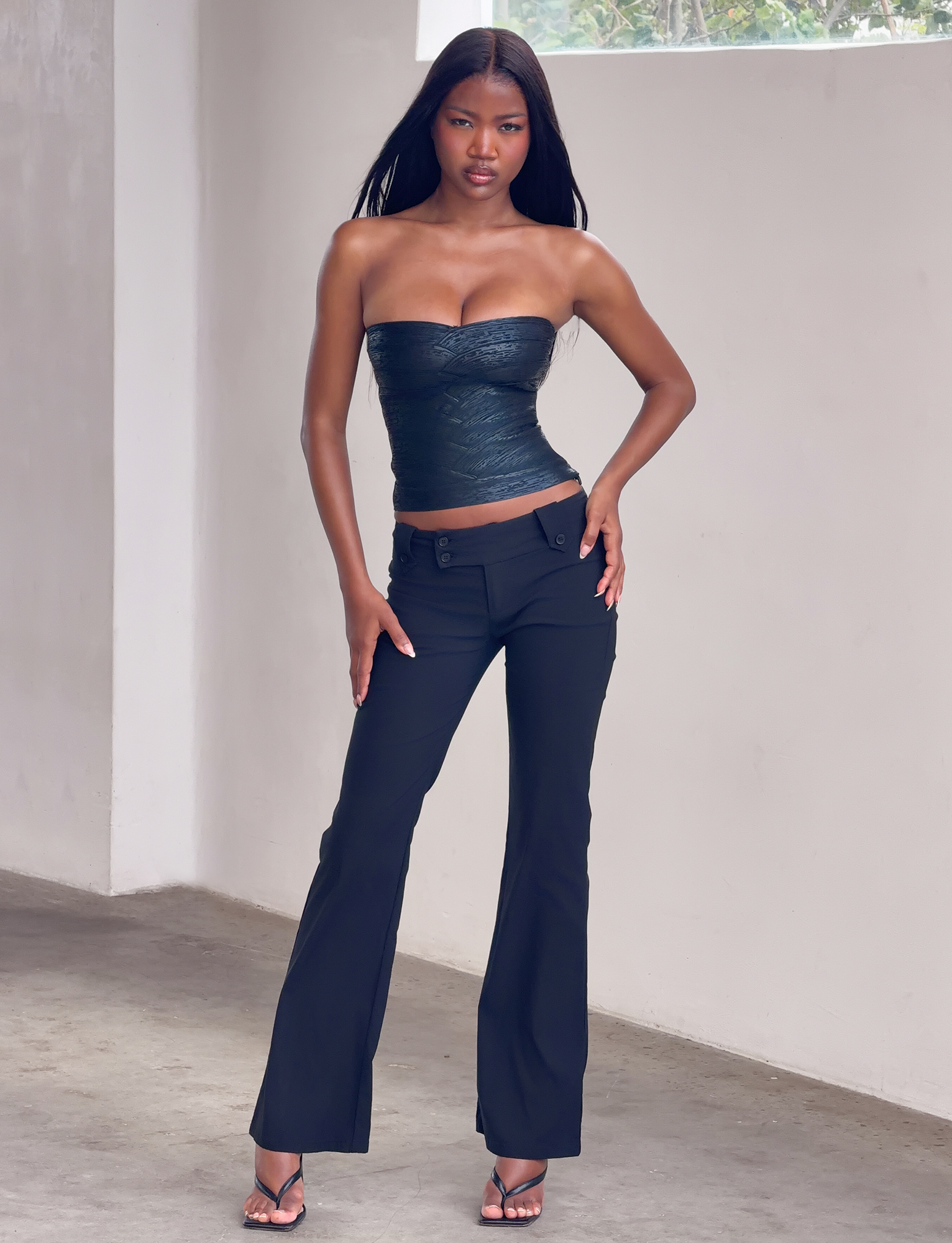 A woman poses indoors against a white wall, wearing the TIGERMIST NIYA METALLIC BANDAGE TOP in black with matching pants and heeled sandals. One hand rests on her hip as she looks confidently at the camera.