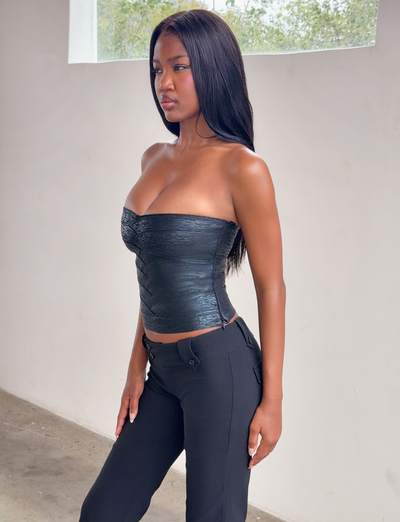 A woman with long straight hair stands indoors by a white wall and window, wearing the TIGERMIST NIYA METALLIC BANDAGE TOP in black and black pants, gazing to the side with a neutral expression.