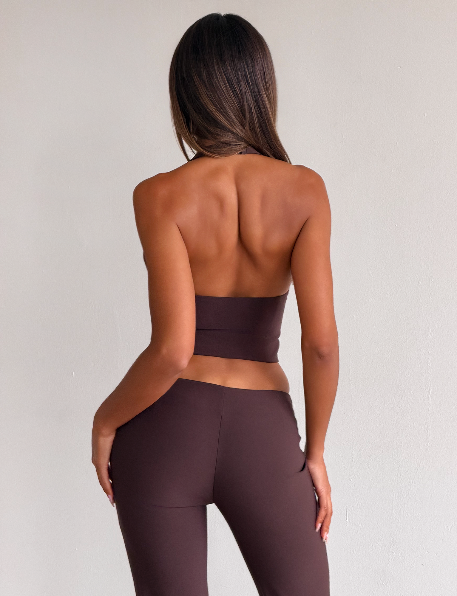 A woman with long hair faces away from the camera, wearing the TIGERMIST KITTIE BUCKLE TOP - CHOCOLATE and matching brown pants against a plain light background.