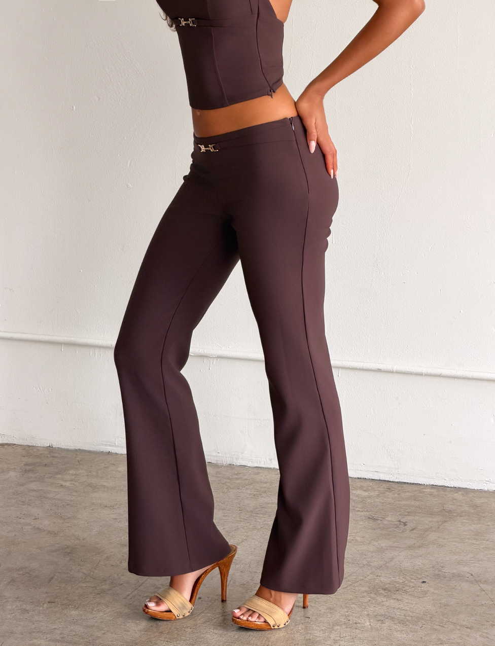 A woman wears the TIGERMIST KITTIE BUCKLE PANT in chocolate, featuring stretch suiting fabric and a flared leg, styled with a matching sleeveless top and tan heels, standing against a white wall with one hand on her hip.