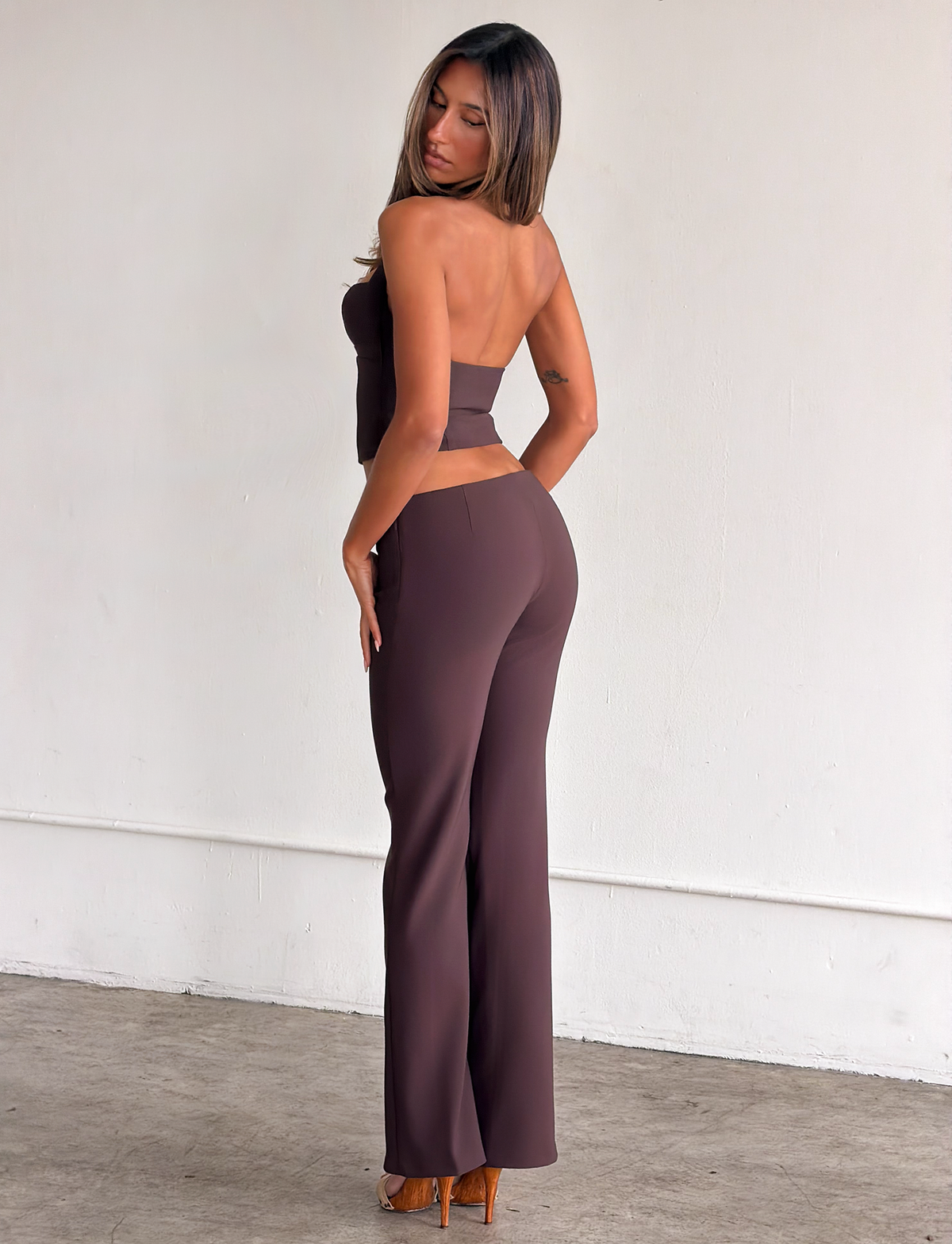 A woman with long hair stands against a plain wall, wearing the TIGERMIST KITTIE BUCKLE PANT in chocolate with flared legs and brown high heels, looking over her shoulder towards the camera.