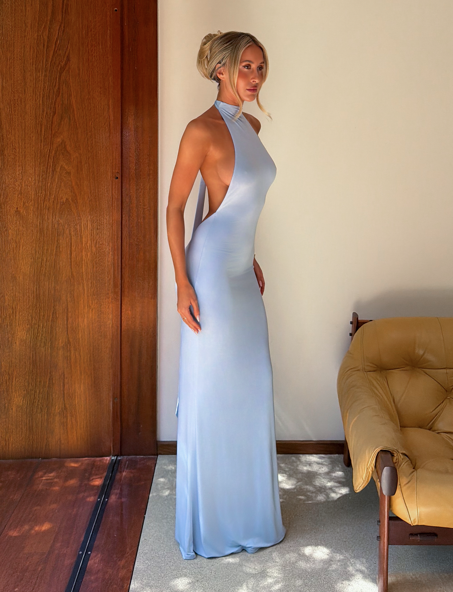 A woman with blonde hair in an updo models the TIGERMIST MADDY MAXI DRESS in baby blue, featuring a halter neck and open back, standing indoors by a wooden wall and beige armchair in soft natural light.