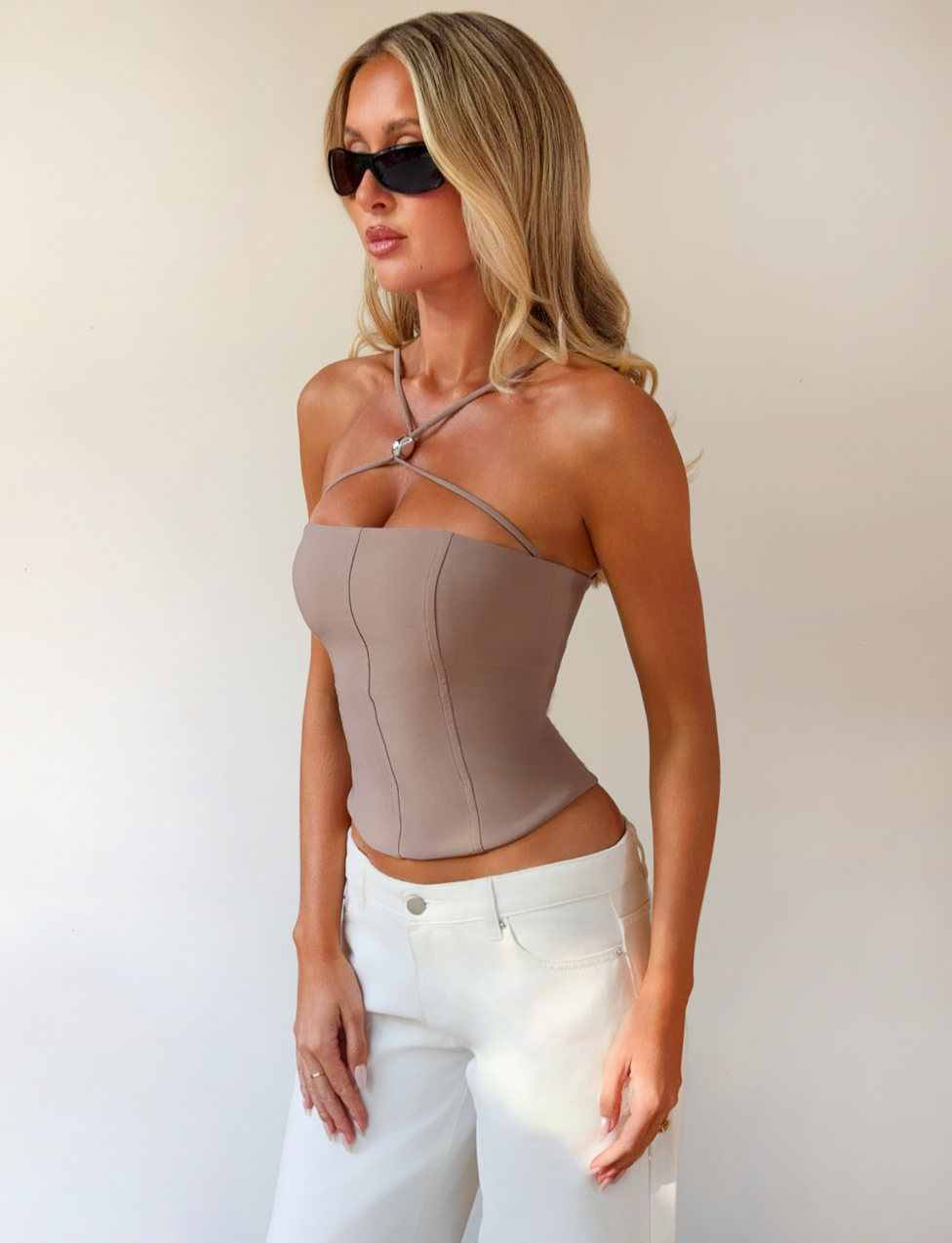 A woman with long blonde hair wears black sunglasses, white pants, and the TIGERMIST TAMORA CORSET TOP in taupe with adjustable straps, standing against a plain light background.