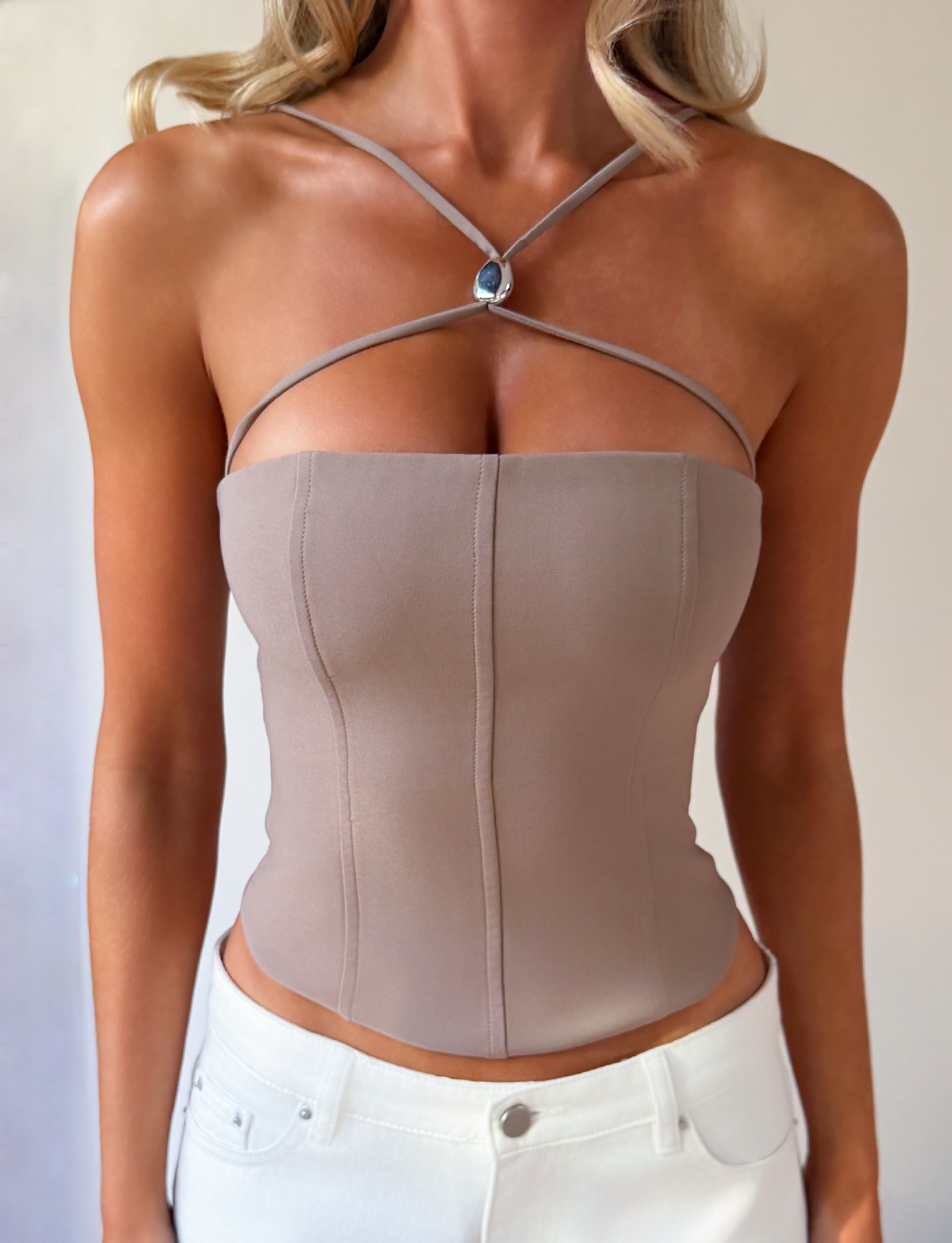 A woman wears the TIGERMIST TAMORA CORSET TOP in taupe with adjustable shoulder straps, styled with a blue gemstone pendant and white pants. Her wavy blonde hair frames her unseen face.