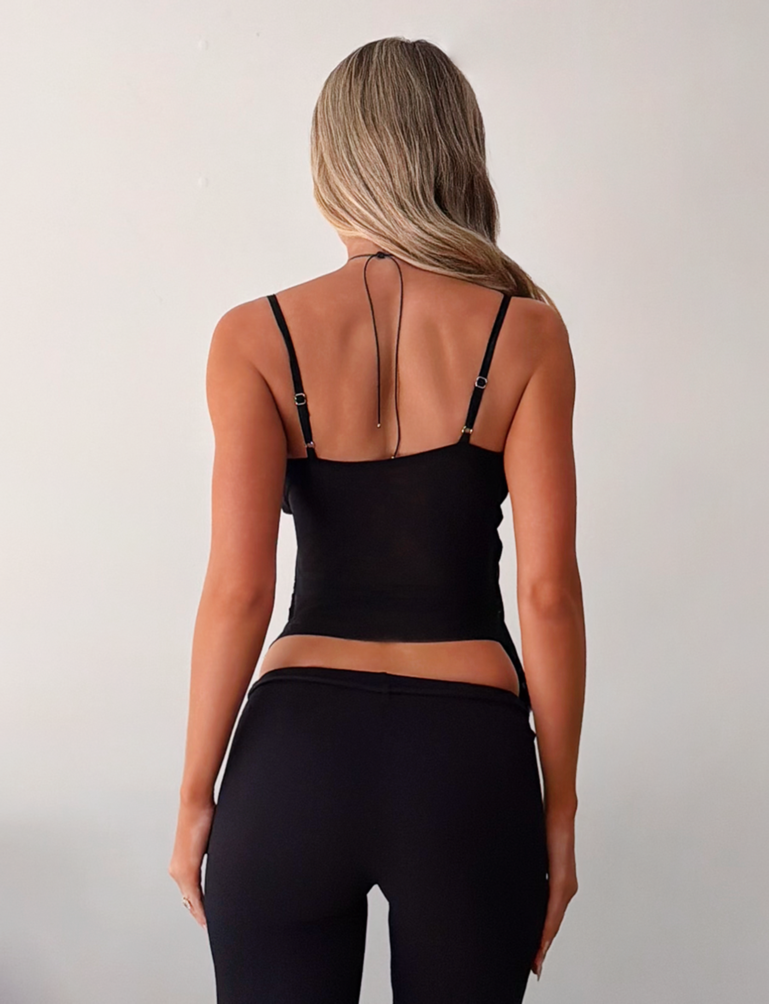 A woman with long, wavy blonde hair faces away, wearing the TIGERMIST ALDEANA TOP in black and black pants against a plain light-colored background.