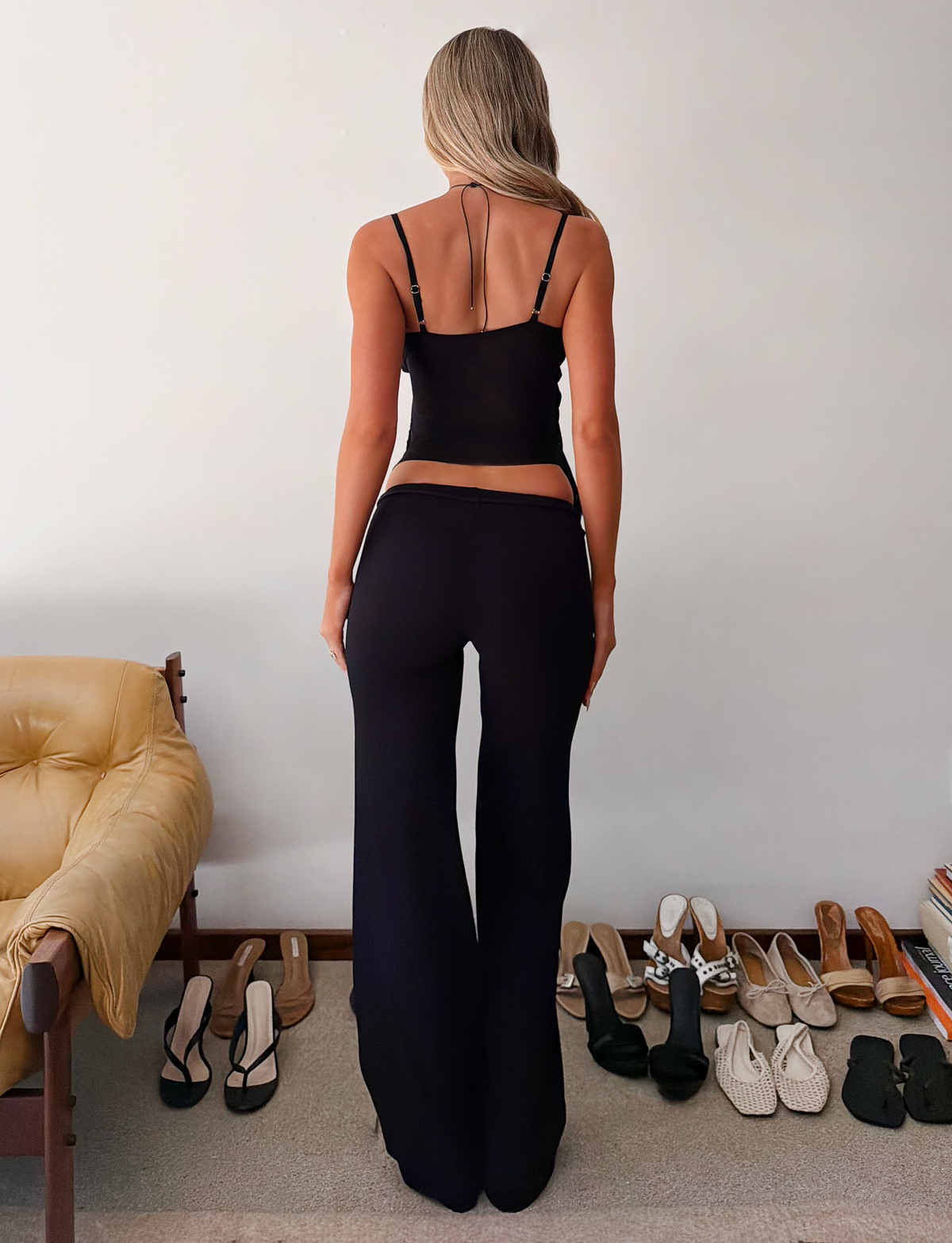A person with long blonde hair, wearing the TIGERMIST KAILI PANT - BLACK and a black sleeveless top, stands facing a white wall. Nearby, several pairs of shoes are lined up beside a beige chair.