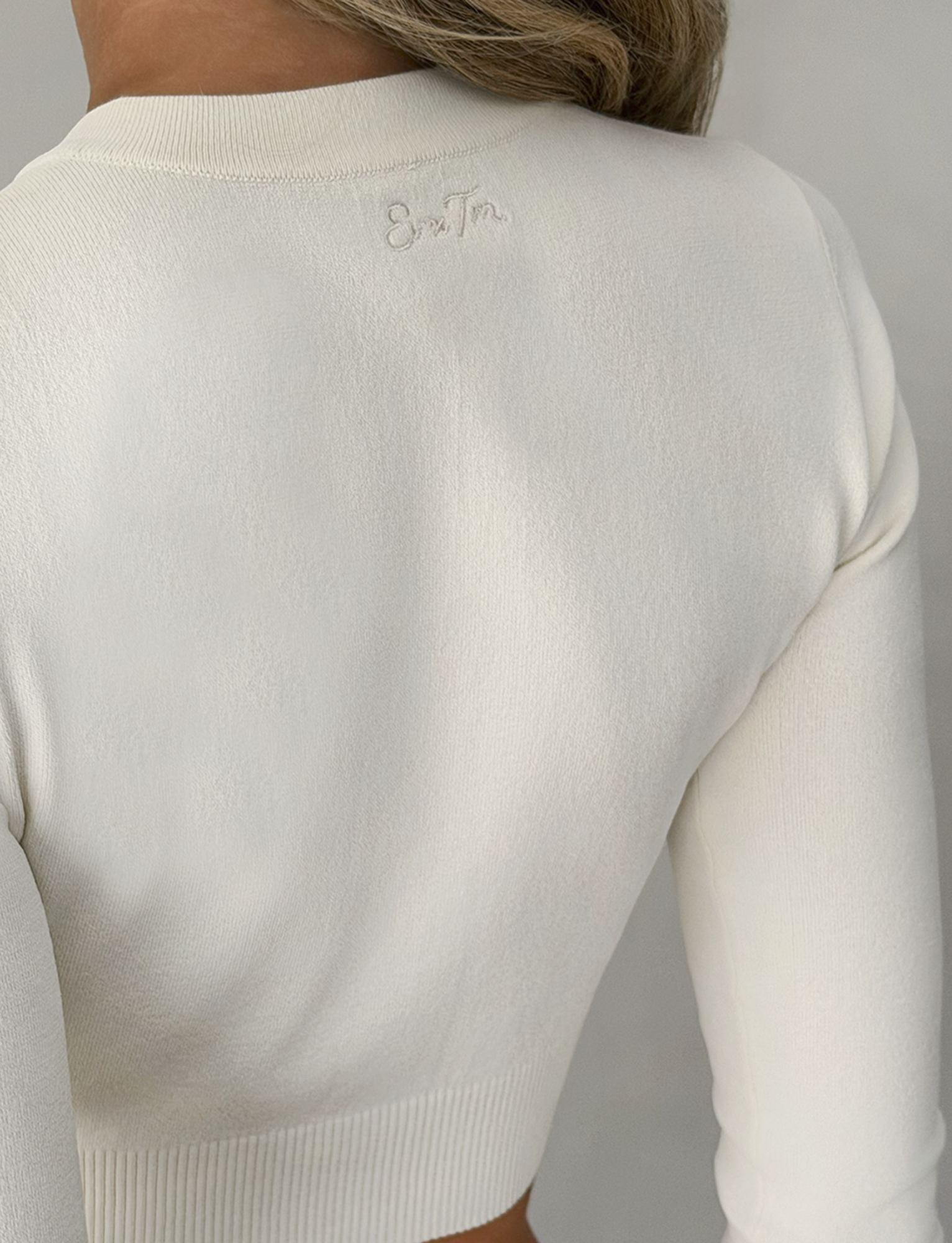 A close-up of a person wearing the TIGERMIST DAMARIS CARDIGAN in cream, showing the back detail with subtle embroidered text near the neckline and hair draped over one shoulder.