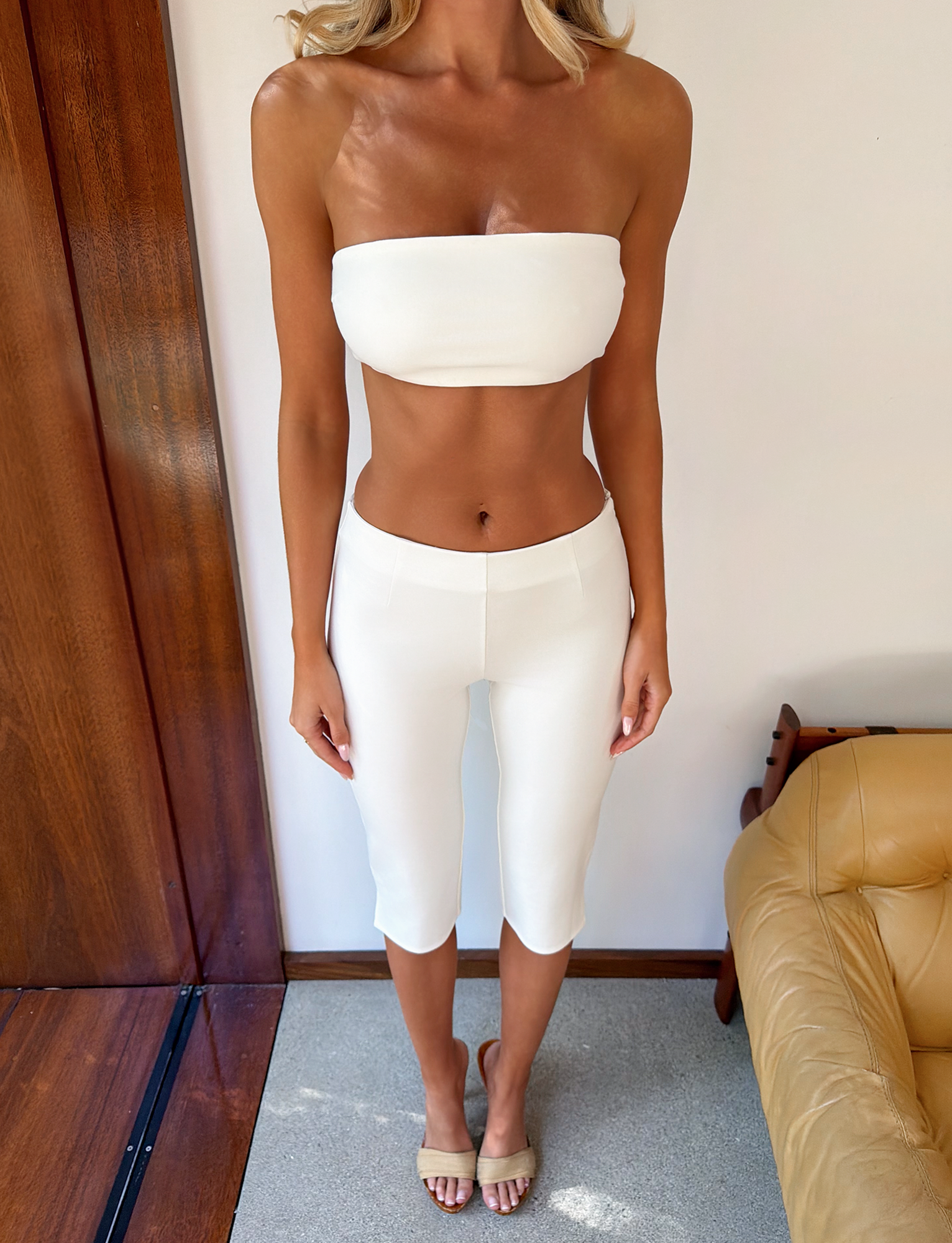 A woman poses indoors wearing the TIGERMIST PILAR BANDEAU in white—a cropped, adjustable lace-up back top—paired with matching high-waisted, knee-length ponte jersey pants. She finishes the look with beige sandals; a tan armchair and wooden wall are behind her.