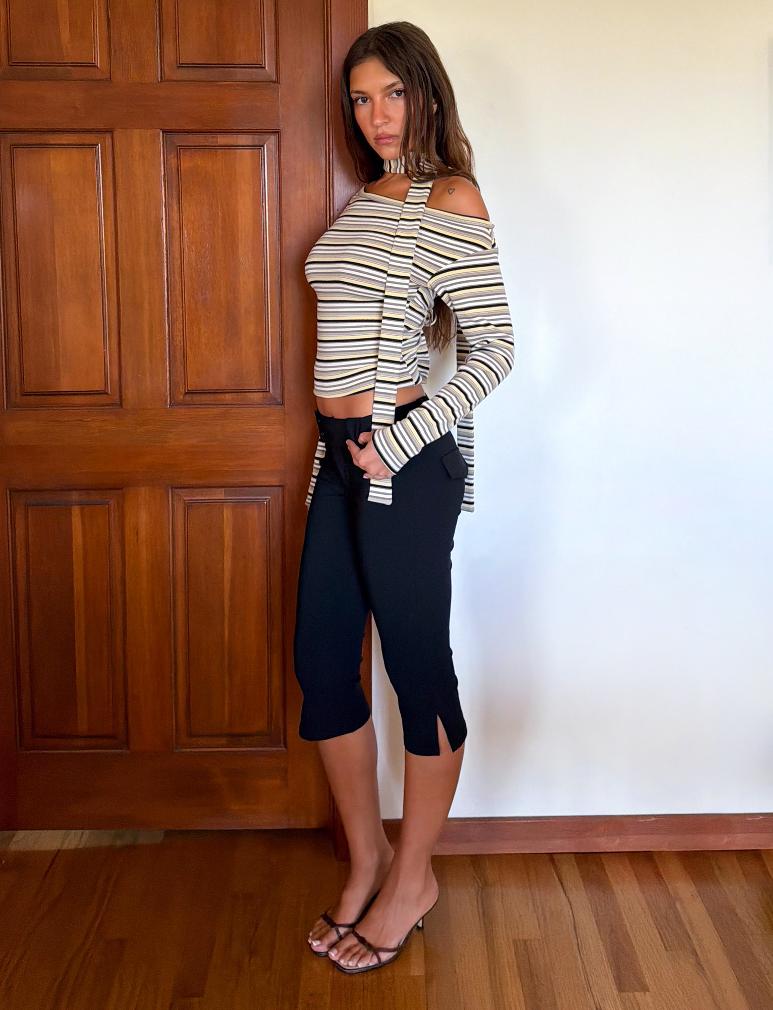 A woman with long brown hair and a neutral expression stands indoors wearing the TIGERMIST KIERSTEN SCARF TOP in MULTISTRIPE, paired with black capri pants and brown strappy sandals.