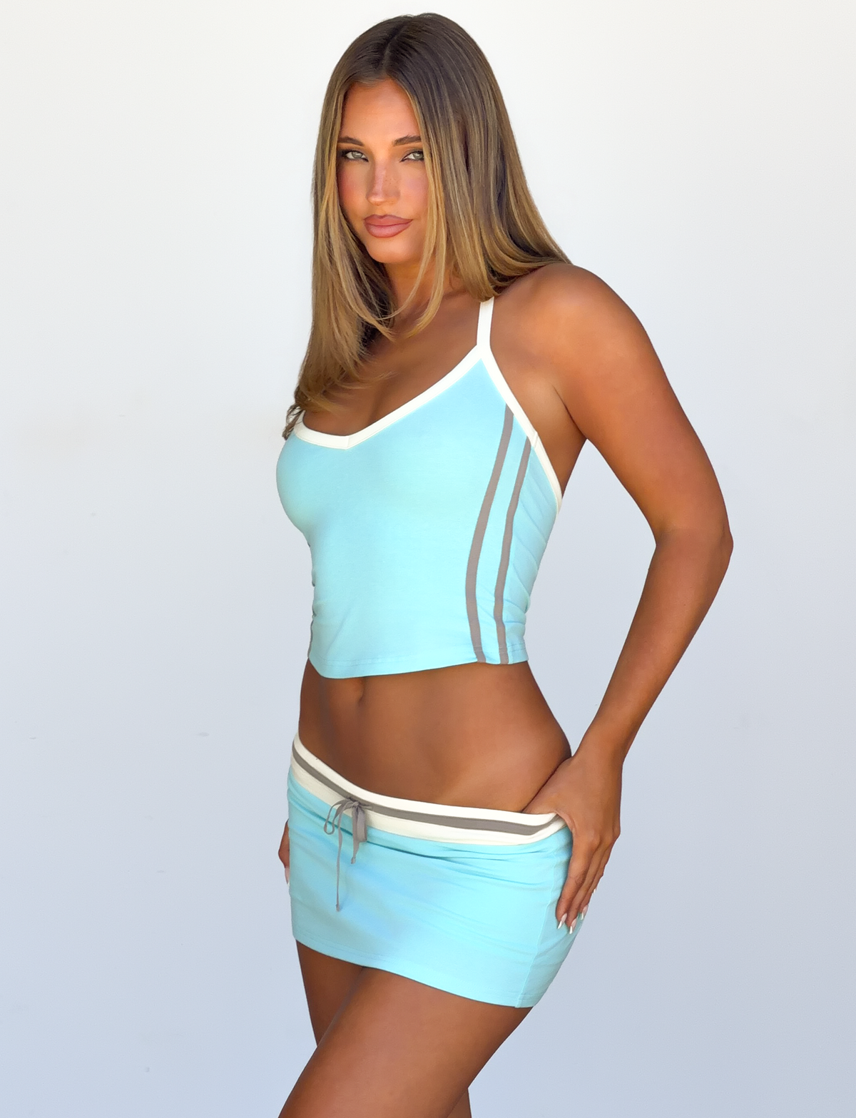 A woman with long hair is wearing the TIGERMIST ALYSIA TOP in aqua, paired with a matching mini skirt featuring contrast binding stripes, as she poses against a plain background with one hand on her hip.