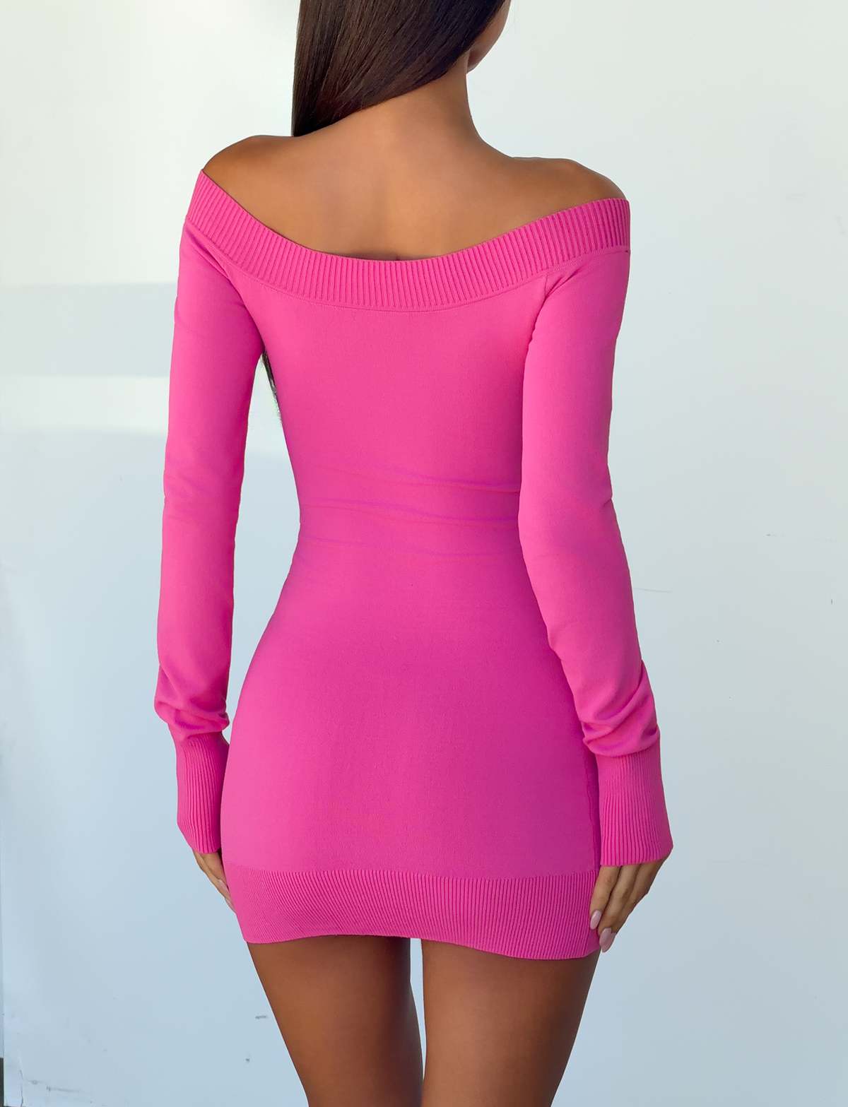 A person with long dark hair is shown from the back, wearing the TIGERMIST PARKER KNIT MINI DRESS in fuchsia, a fitted, off-the-shoulder style with long sleeves. The background is plain and light-colored.