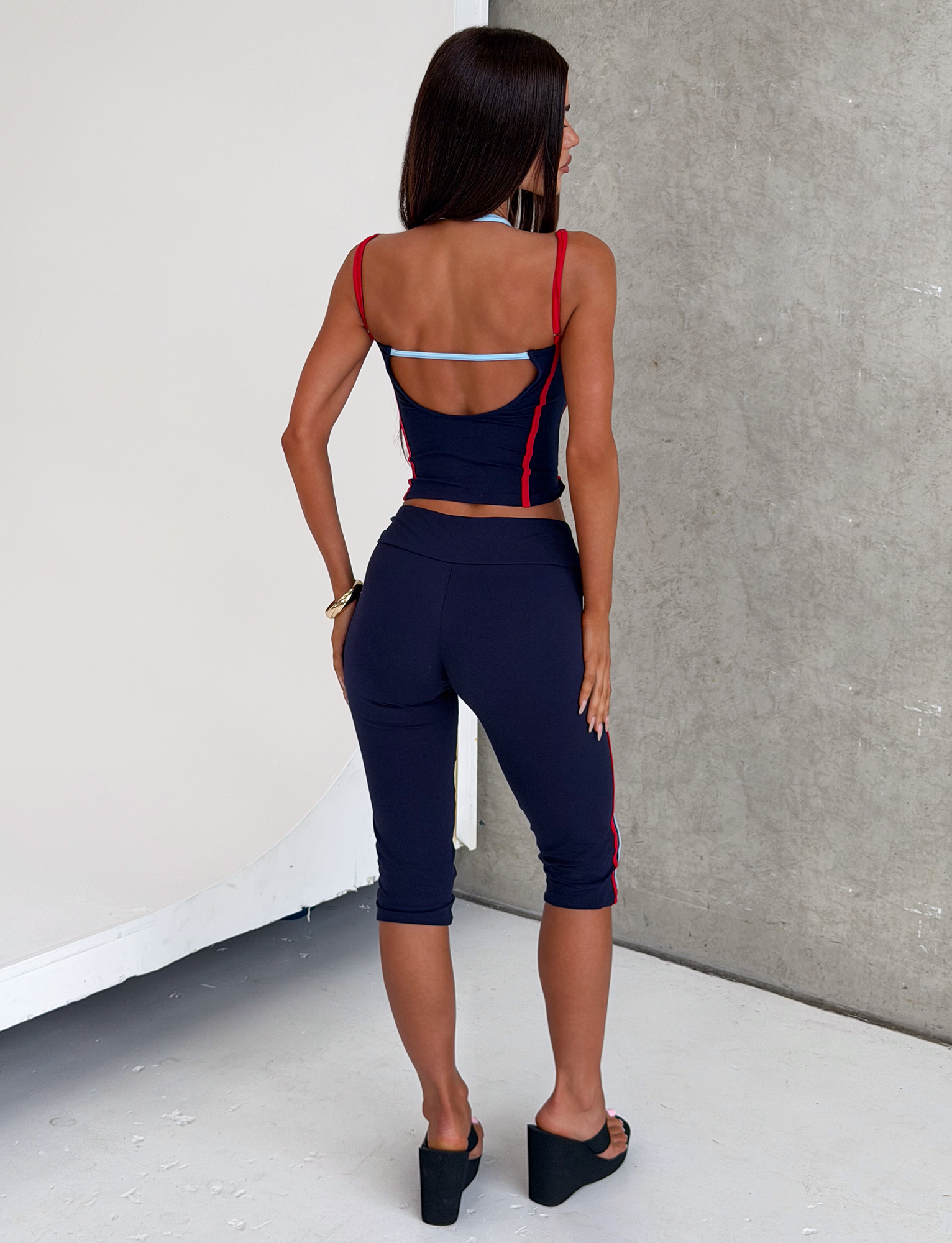 A woman with long, straight hair wears the TIGERMIST TATUM CAPRI - NAVY STRIPE set: a navy crop top with red trim and matching striped capris. She pairs it with black platform sandals and stands before a gray textured wall.