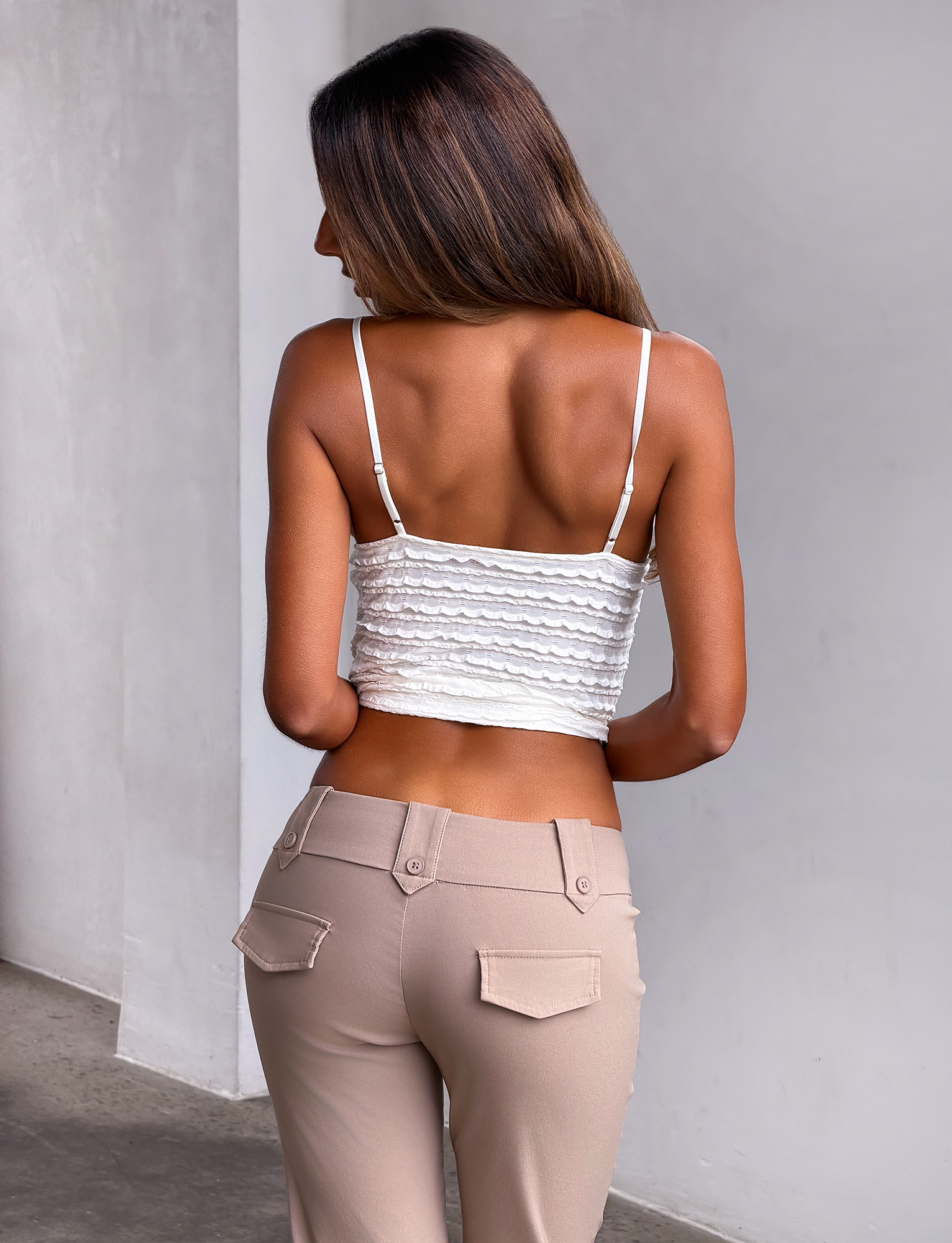 A woman with long brown hair stands with her back to the camera, wearing the TIGERMIST ZORA TOP in cream, featuring adjustable shoulder straps, paired with high-waisted beige pants against a plain light wall and floor.
