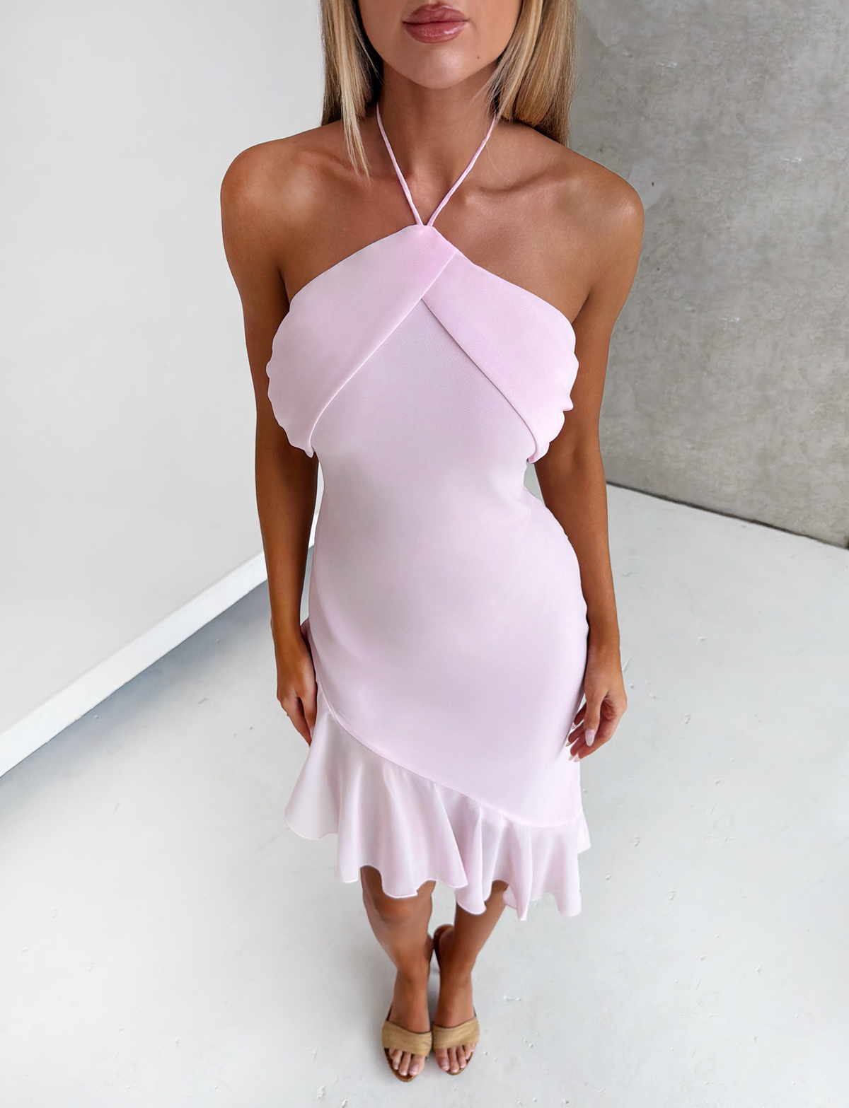 A woman stands indoors on a light floor wearing the TIGERMIST ELARINA MIDI DRESS in pink, featuring a halter-neck, ruffle details, and an asymmetrical hemline. Her hands are relaxed by her sides and her face is partially out of frame.