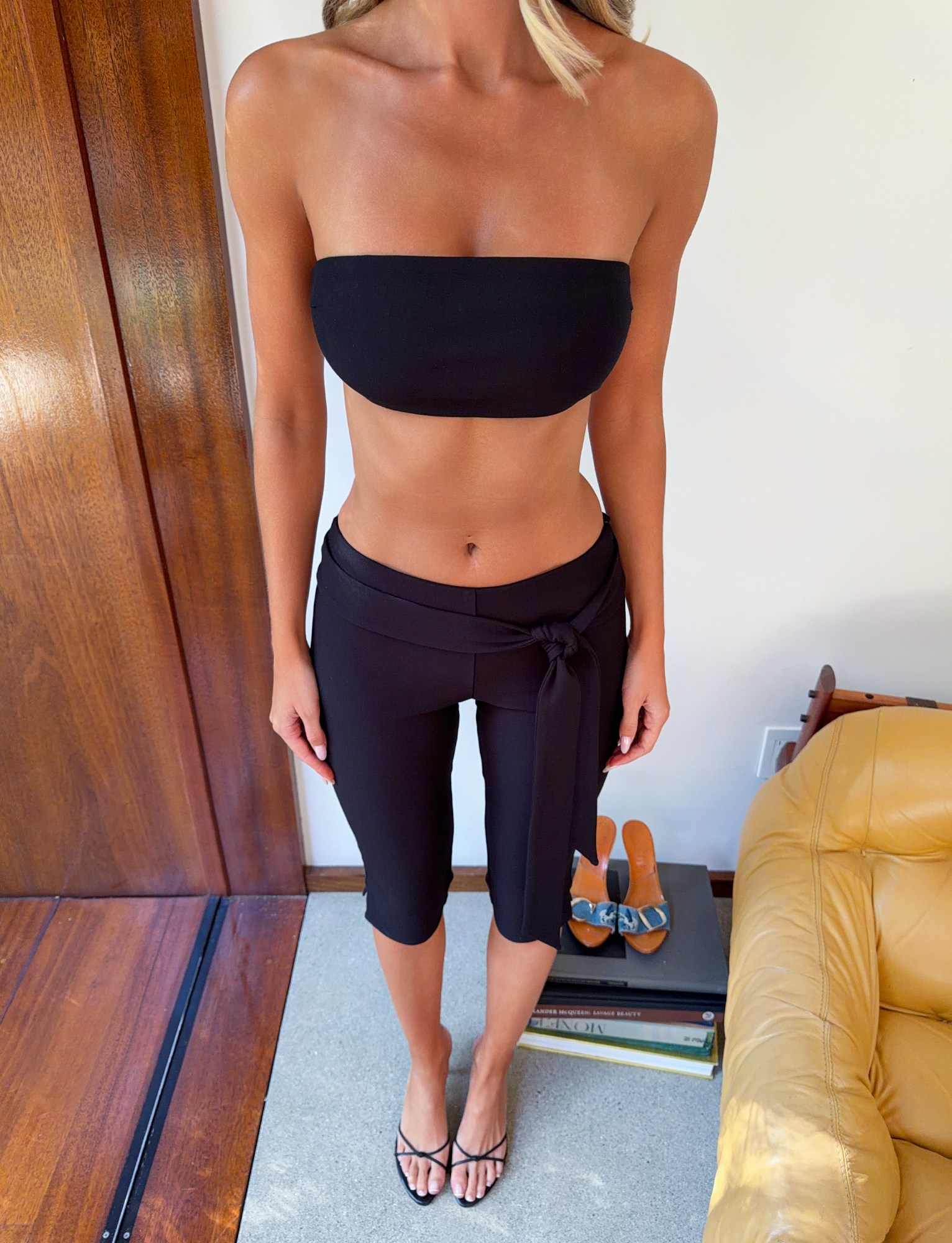 A woman stands indoors wearing the TIGERMIST PILAR BANDEAU in black, paired with black wrap-style ponte jersey pants and sandals. Her arms are relaxed by her sides amid a chair, books, and extra sandals on the floor.