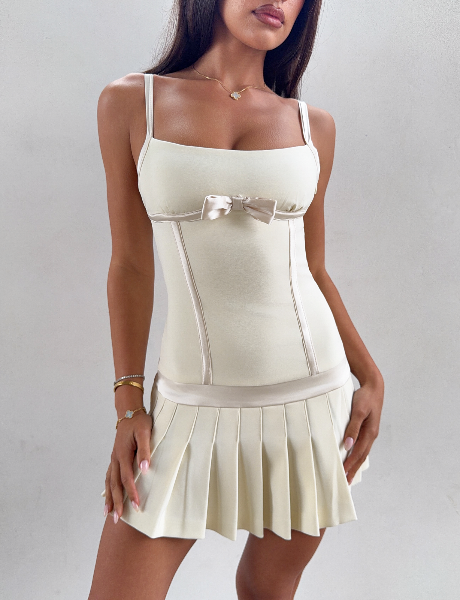 A woman models the TIGERMIST MARLOW MINI DRESS in cream—a sleeveless, pleated mini dress featuring a small bow on the bodice and a satin waistband—while standing against a plain light background.