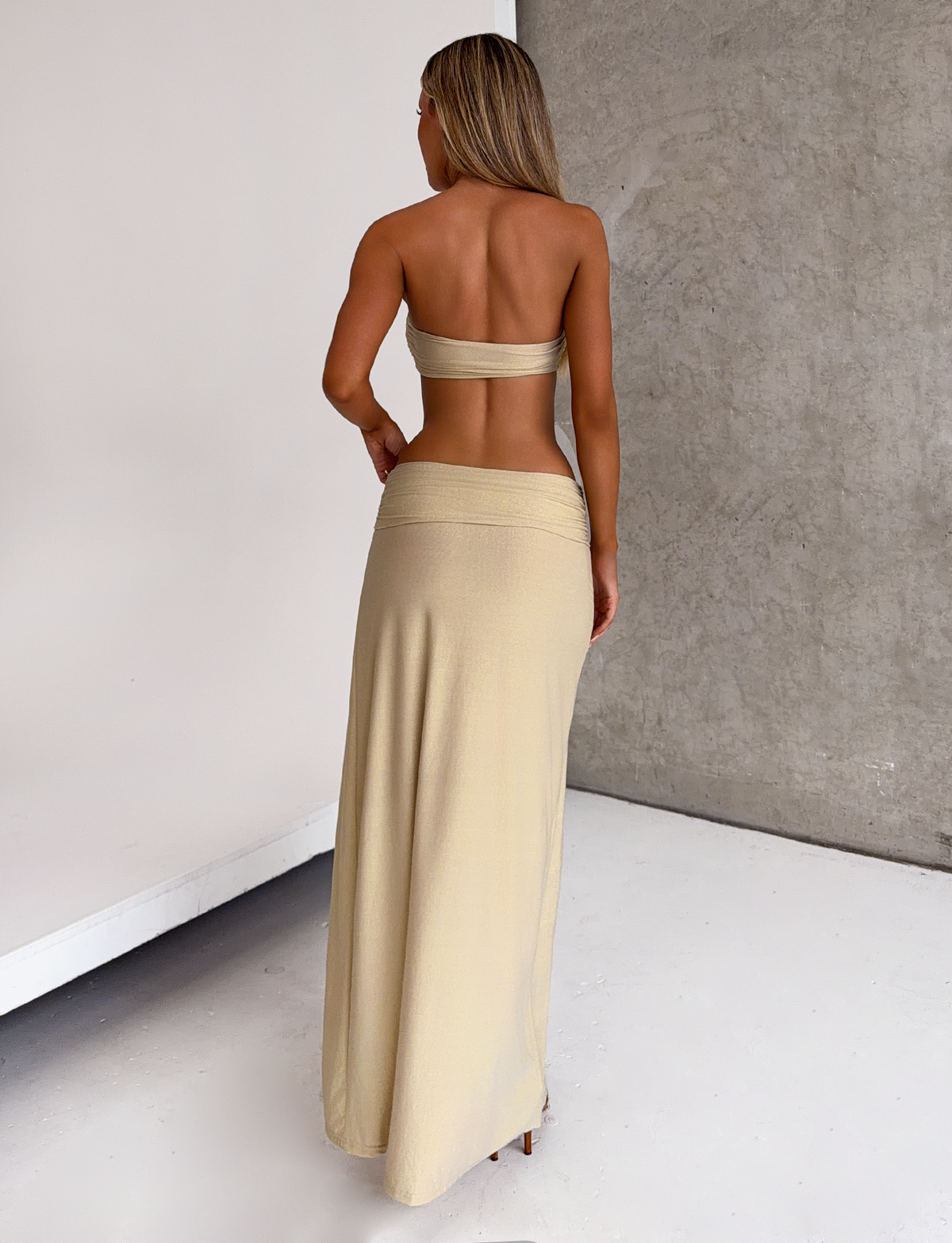 A woman stands with her back to the camera, wearing the TIGERMIST KYNLEE 2.0 MAXI DRESS in Gold, in a minimalist indoor space with a concrete wall and white floor.