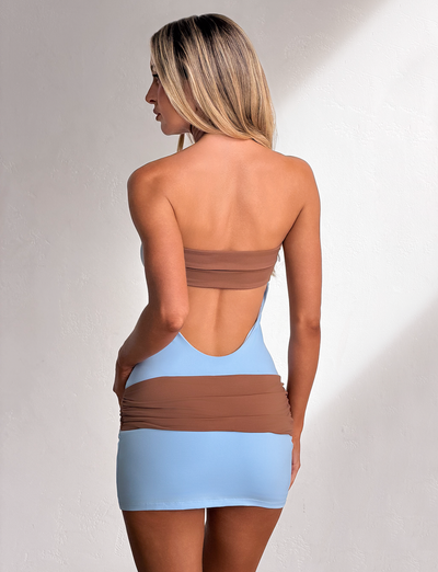 A woman with long blonde hair models the TIGERMIST EOS MINI DRESS in soft blue and mocha, standing against a light textured wall, facing away and glancing to the side.