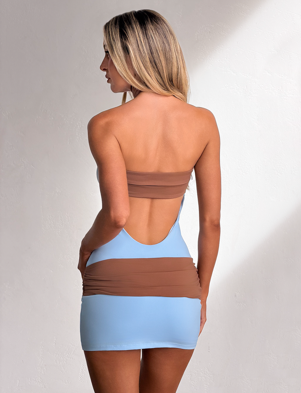 A woman with long blonde hair models the TIGERMIST EOS MINI DRESS in soft blue and mocha, standing against a light textured wall, facing away and glancing to the side.