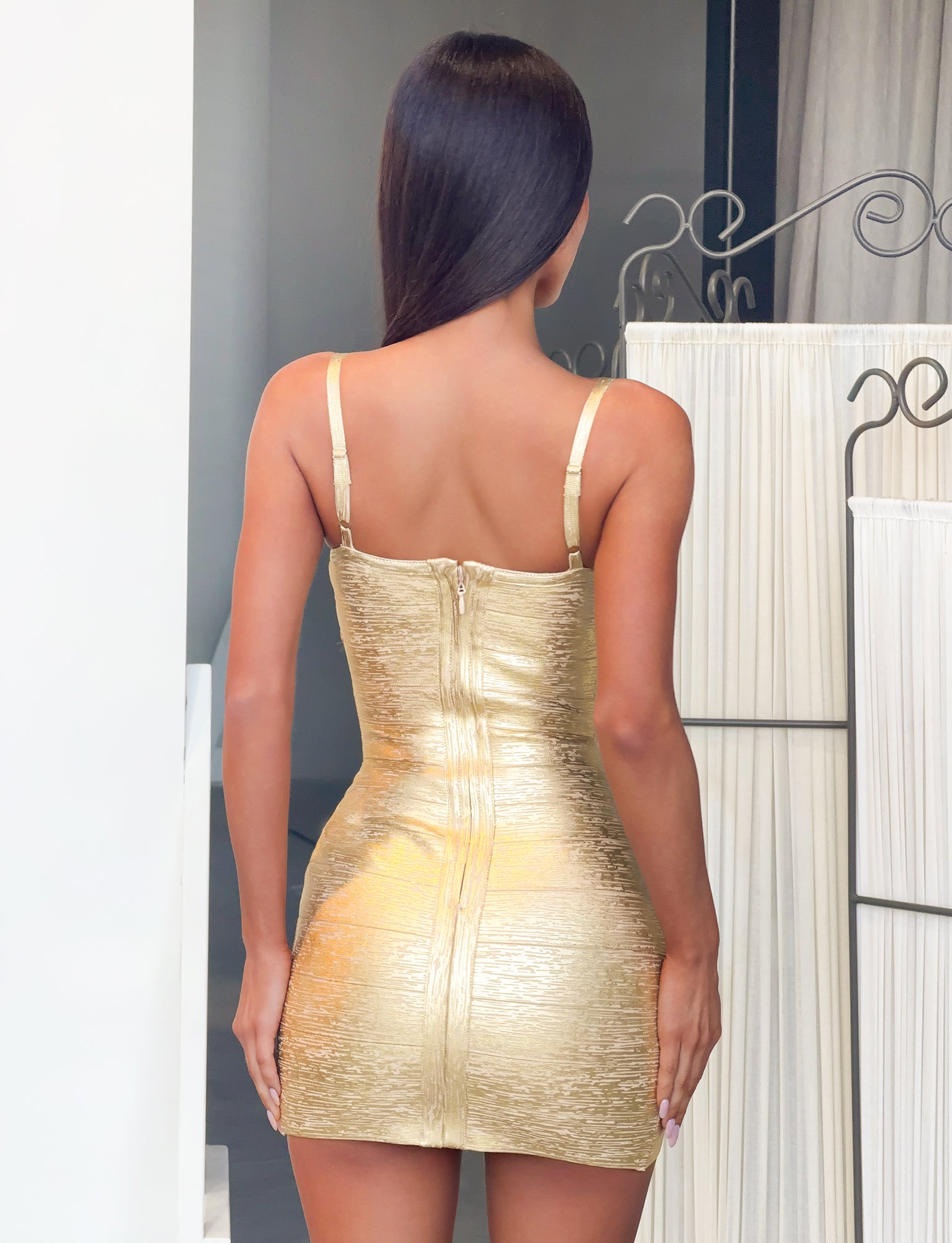 A woman with long dark hair stands facing away, wearing the TIGERMIST KENZA BANDAGE MINI DRESS in metallic gold. This shimmer-textured, bodycon mini dress features thin straps and a prominent center zipper, making it an ideal choice for special occasions. She is indoors near white walls and a decorative divider.