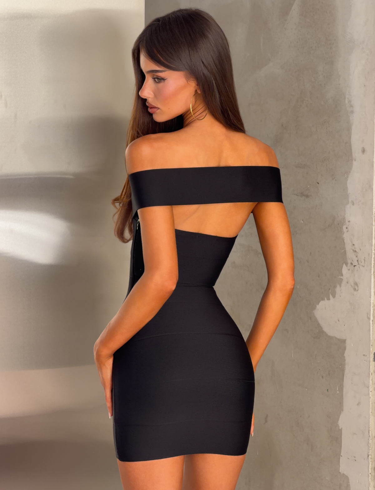 A woman with long brown hair poses sideways, wearing the TIGERMIST ANTONEA BANDAGE MINI DRESS in black, which features an off-shoulder, form-fitting silhouette, as she stands against a textured beige and metallic background.