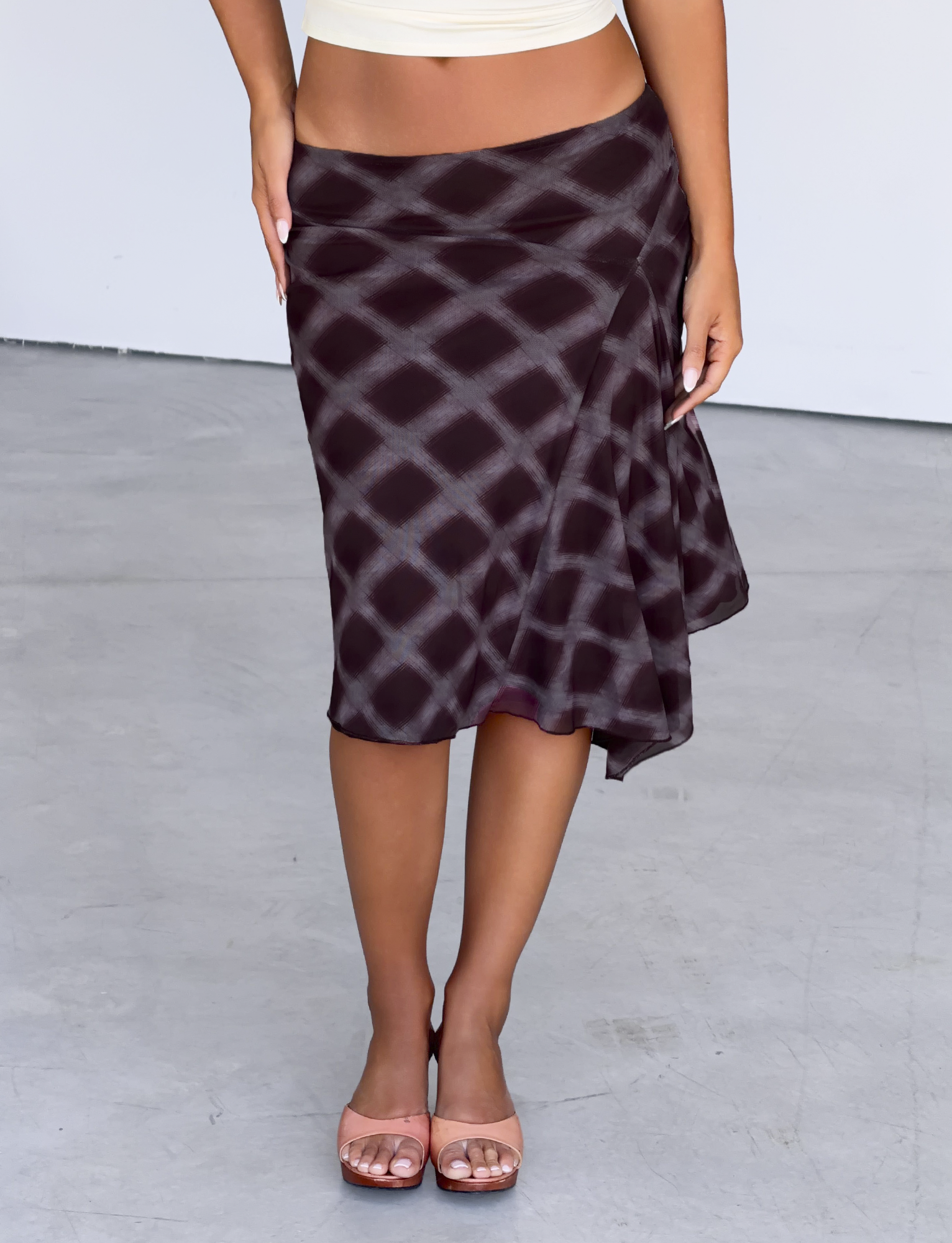 A person wearing the TIGERMIST SAFFRON MIDI SKIRT in Mahogany Check, styled low-waisted with nude heels, stands on a light gray floor, their upper body mostly out of frame.