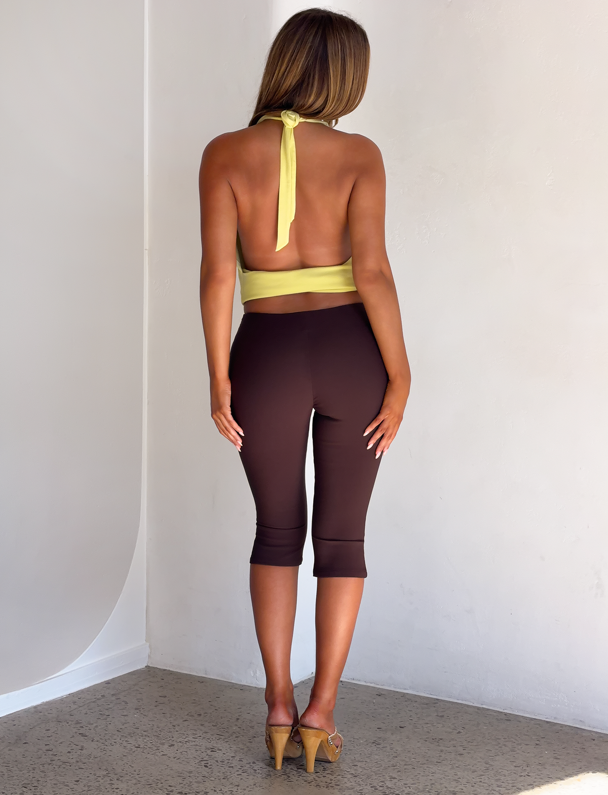 A person seen from behind wearing a pistachio green LYDIAH TOP - PISTACHIO halter neck top, dark brown cropped leggings, and heeled sandals.