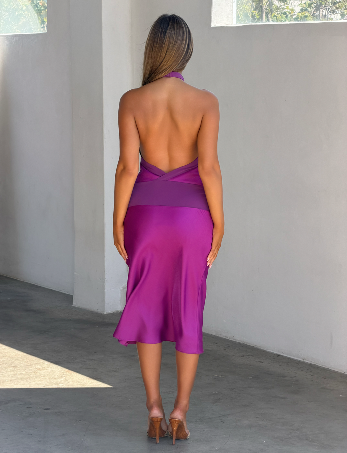 A woman stands indoors facing away, wearing the TIGERMIST ZAYA MIDI DRESS - MULBERRY and tan heels. Sunlight from above casts soft shadows on the light gray floor and wall.