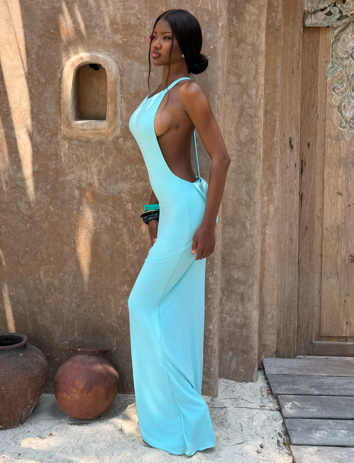 A woman in the RONI MAXI DRESS - AQUA by TIGERMIST stands sideways on sandy ground by rustic clay pots and a textured brown wall with carved wood accents.