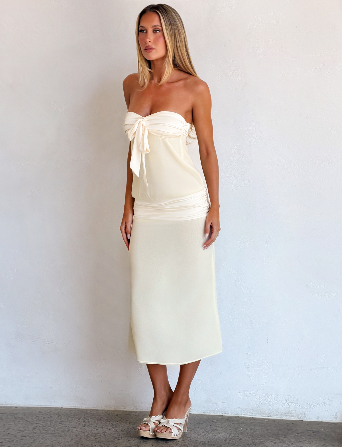 A woman wearing a TIGERMIST LUX MIDI DRESS - CREAM, a strapless cream satin midi dress with a tied bodice detail, and heeled sandals.