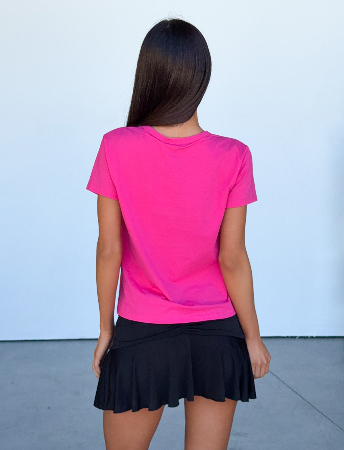 A woman with long brown hair stands facing away against a plain light background, wearing the TIGERMIST BELLA BABY TEE in hot pink with a crew neckline paired with a black pleated skirt.