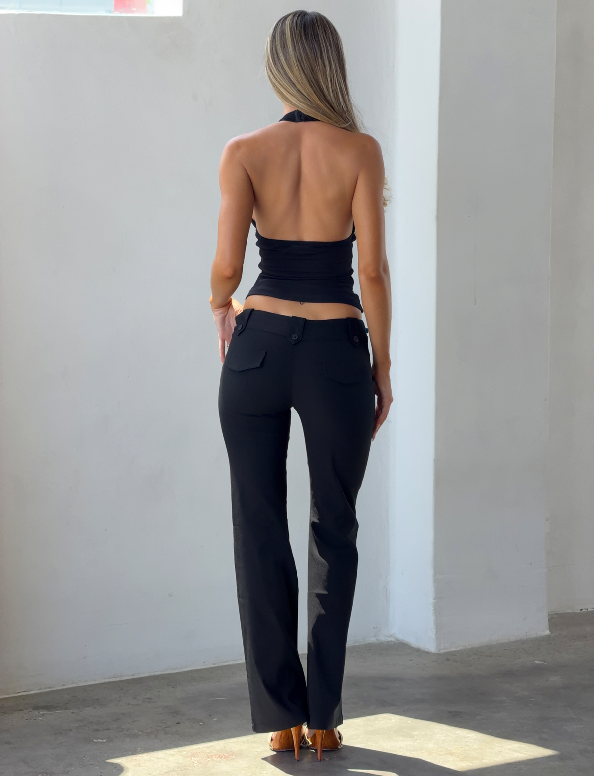 A person viewed from behind, wearing a black NINAH TOP - BLACK mesh halter top and black trousers.
