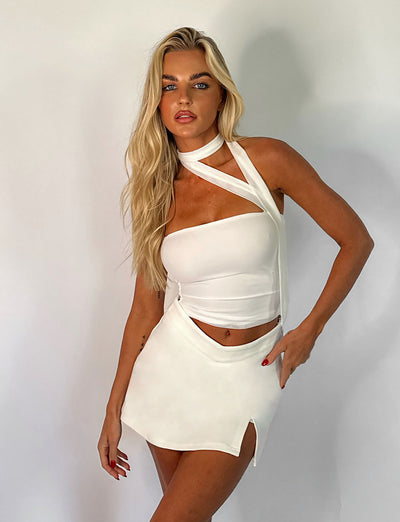 A woman with long blonde hair poses against a white background, wearing a white asymmetrical sleeveless top paired with the DAPHNIE SKORT - WHITE from TIGERMIST. The mini-length skort highlights her red nails as she gazes confidently at the camera.