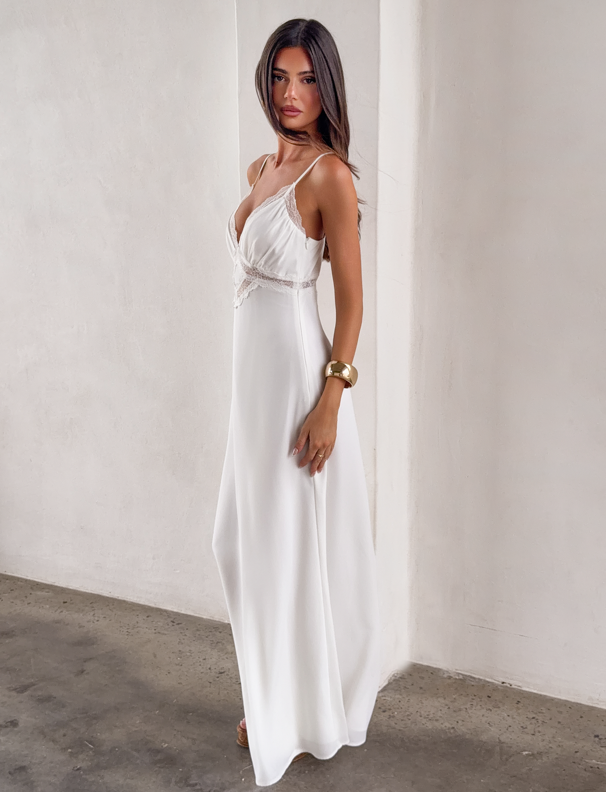 A woman poses confidently in the TIGERMIST AISLYNN MAXI DRESS - WHITE, featuring lace details. She accessorizes with a gold bracelet and has long, straight hair, standing against a textured light-colored wall.