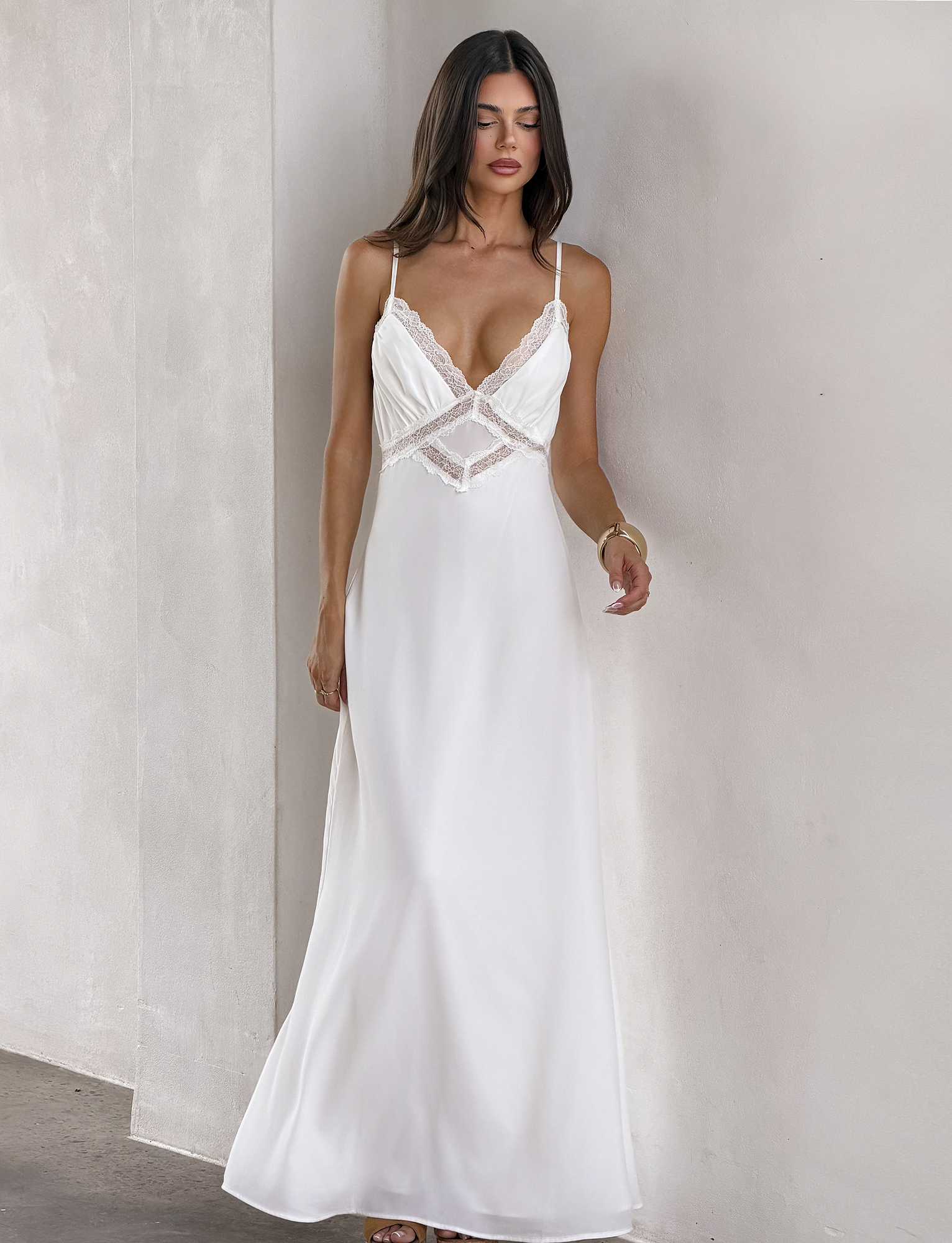 A woman with loose hair and a neutral expression stands against a light wall, wearing the TIGERMIST AISLYNN MAXI DRESS - WHITE—a long slip dress featuring lace trim and thin straps.