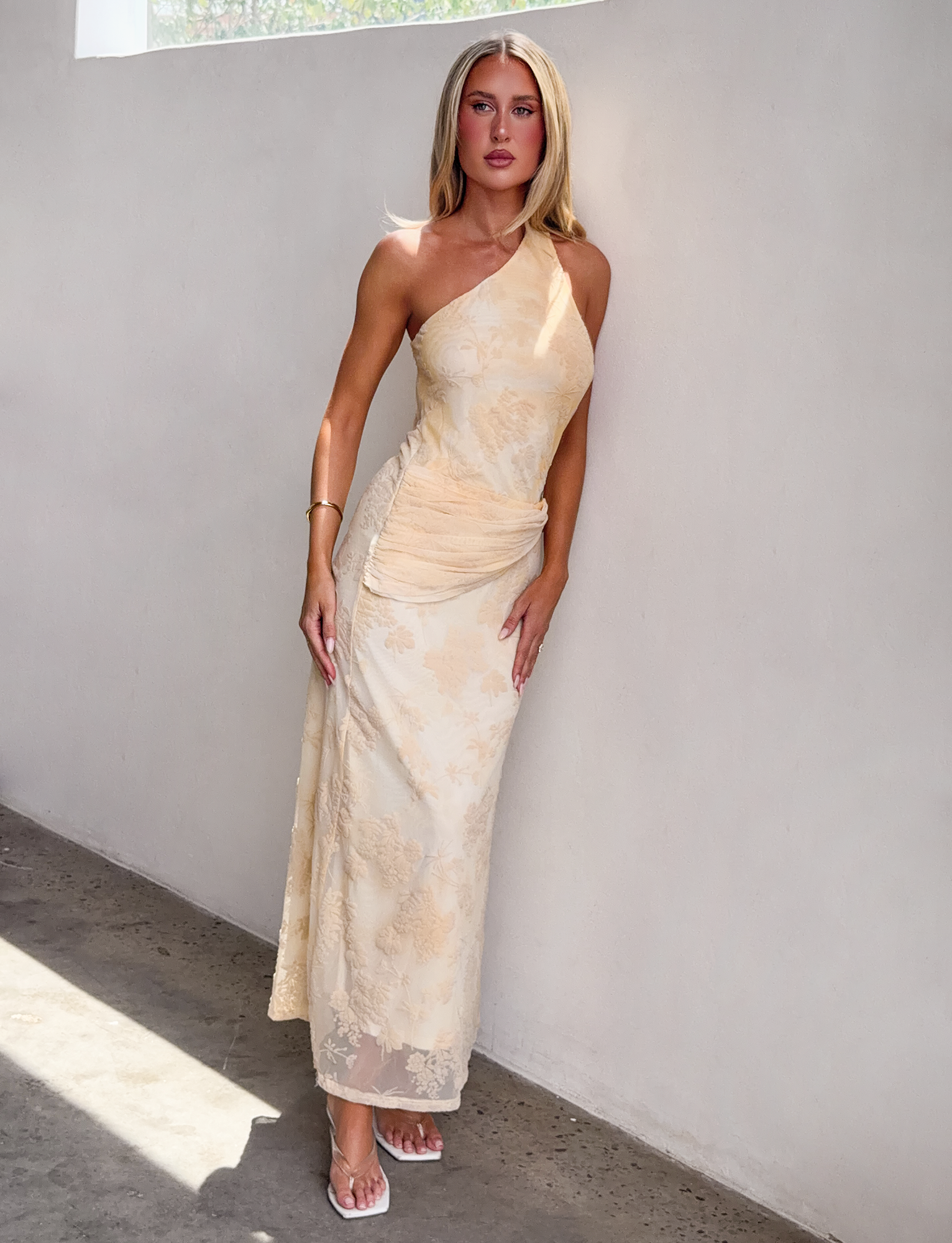 A woman stands indoors by a light wall, wearing the TIGERMIST CHARLYSE ONE SHOULDER MAXI DRESS in butter yellow. She has long blonde hair and heeled sandals, with sunlight streaming in from the left.