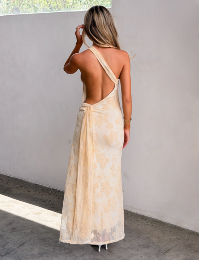 A woman with light brown hair and white heels stands indoors by a light wall, wearing the TIGERMIST CHARLYSE ONE SHOULDER MAXI DRESS in butter yellow, featuring floral mesh fabric and a chic ruched waistband.