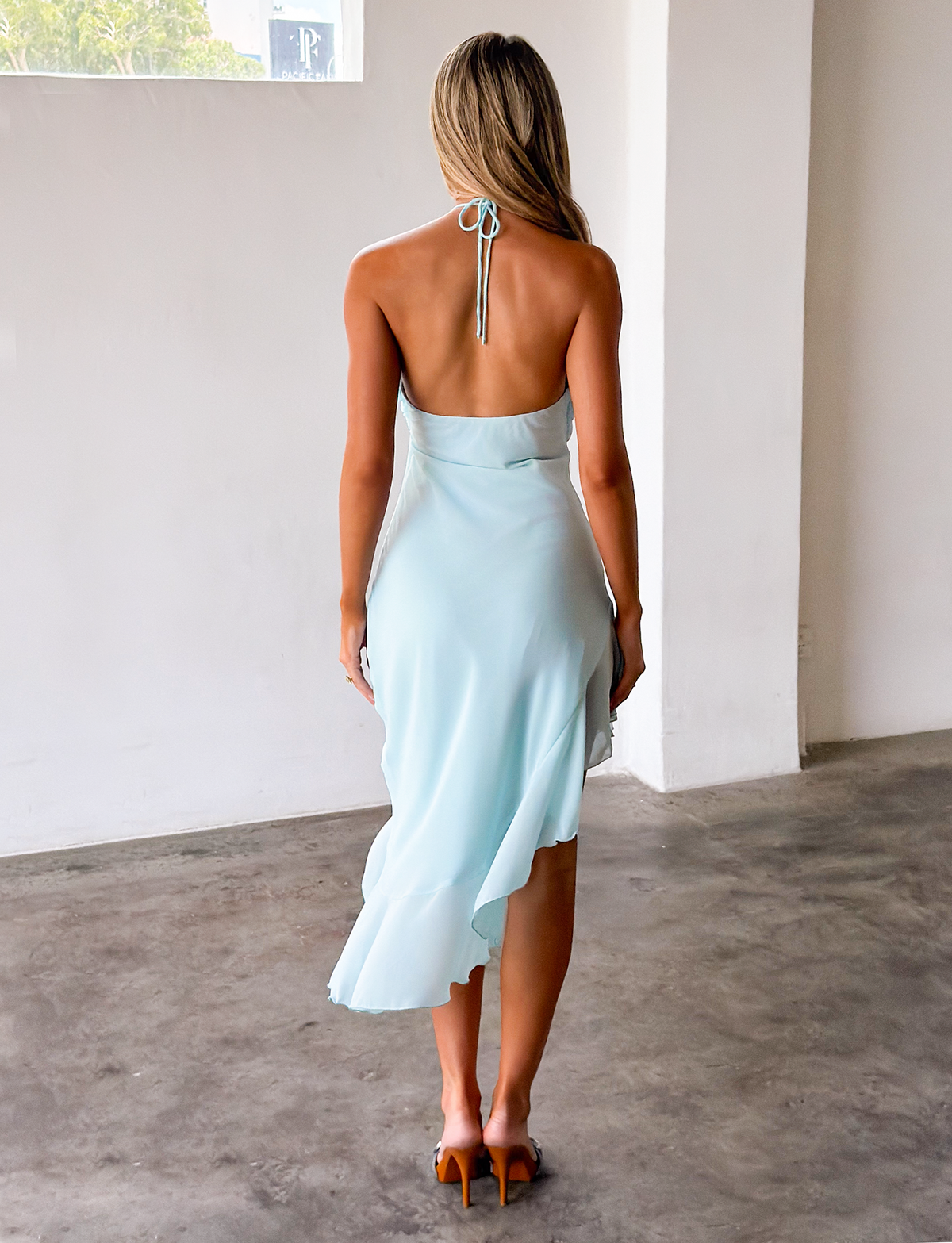 A woman with long blonde hair stands indoors on a concrete floor, facing away from the camera, wearing the TIGERMIST ELARINA MIDI DRESS in Mermaid Blue and brown heeled sandals.
