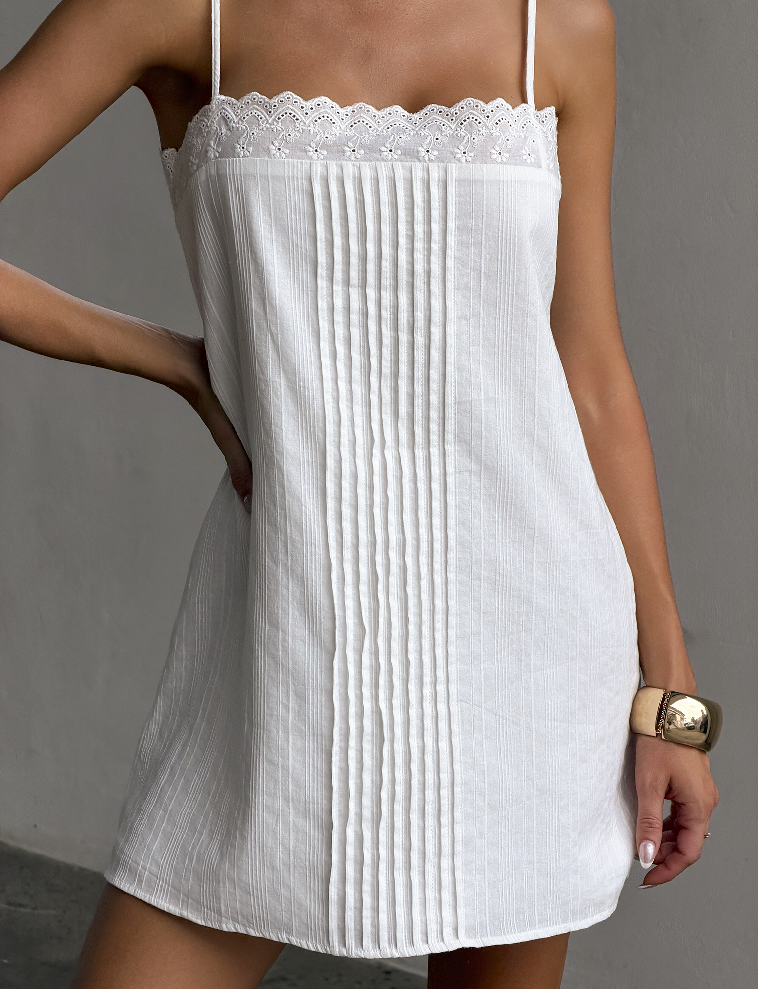 Wearing the TIGERMIST BRYNLEE MINI DRESS in white, featuring adjustable straps, lace-trimmed neckline, and front pleats, a person poses with one hand on their hip and pairs it with a chunky gold bracelet.