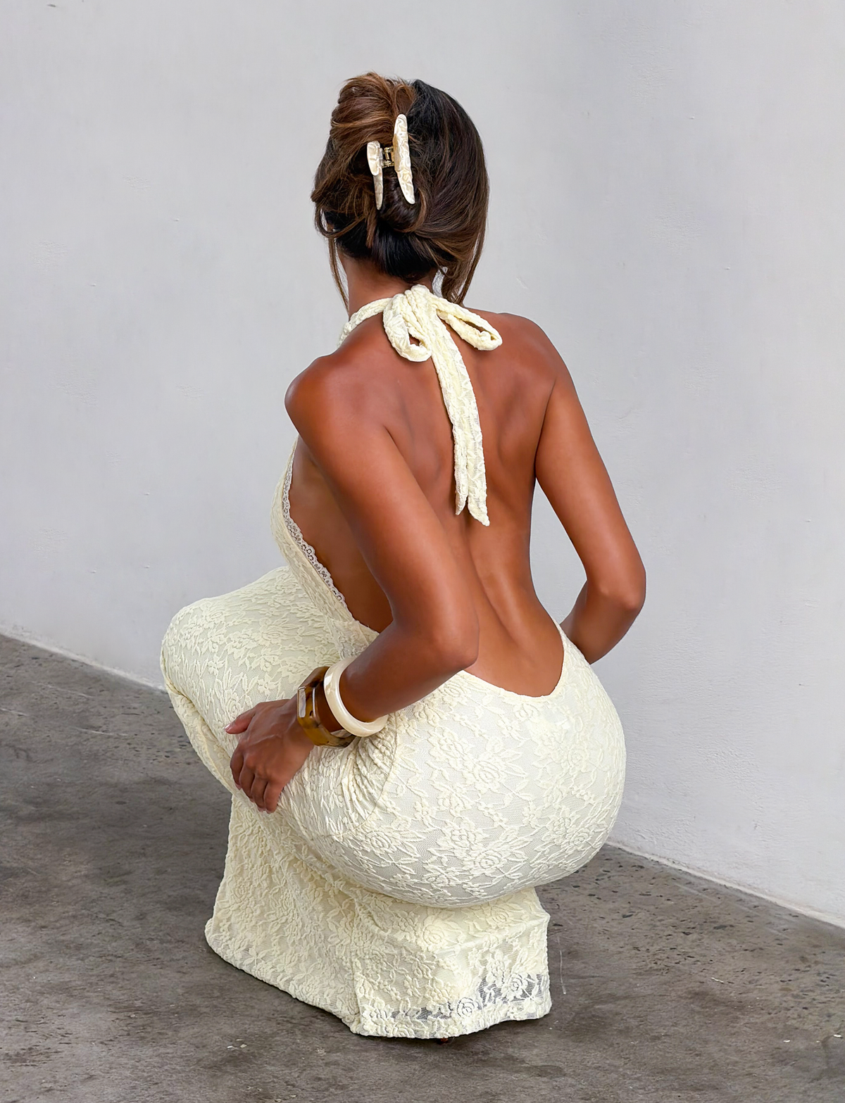 A woman with her back to the camera squats near a white wall, wearing the TIGERMIST MAVEN LACE MAXI DRESS in pastel yellow with matching hair accessories, white bangles on her right wrist, and a delicate halter neckline.