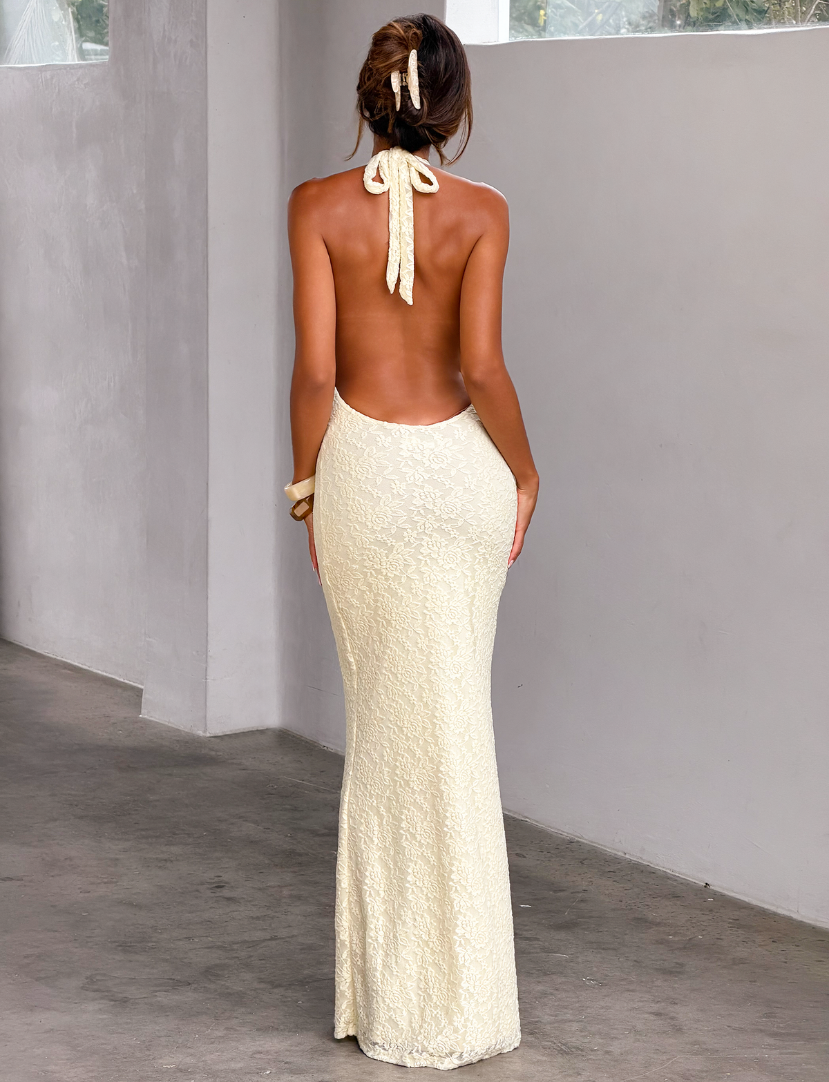 A woman stands indoors with her back turned, wearing the TIGERMIST MAVEN LACE MAXI DRESS in pastel yellow. Her hair is tied with a matching ribbon as she stands by a gray wall and window, hands relaxed at her sides.